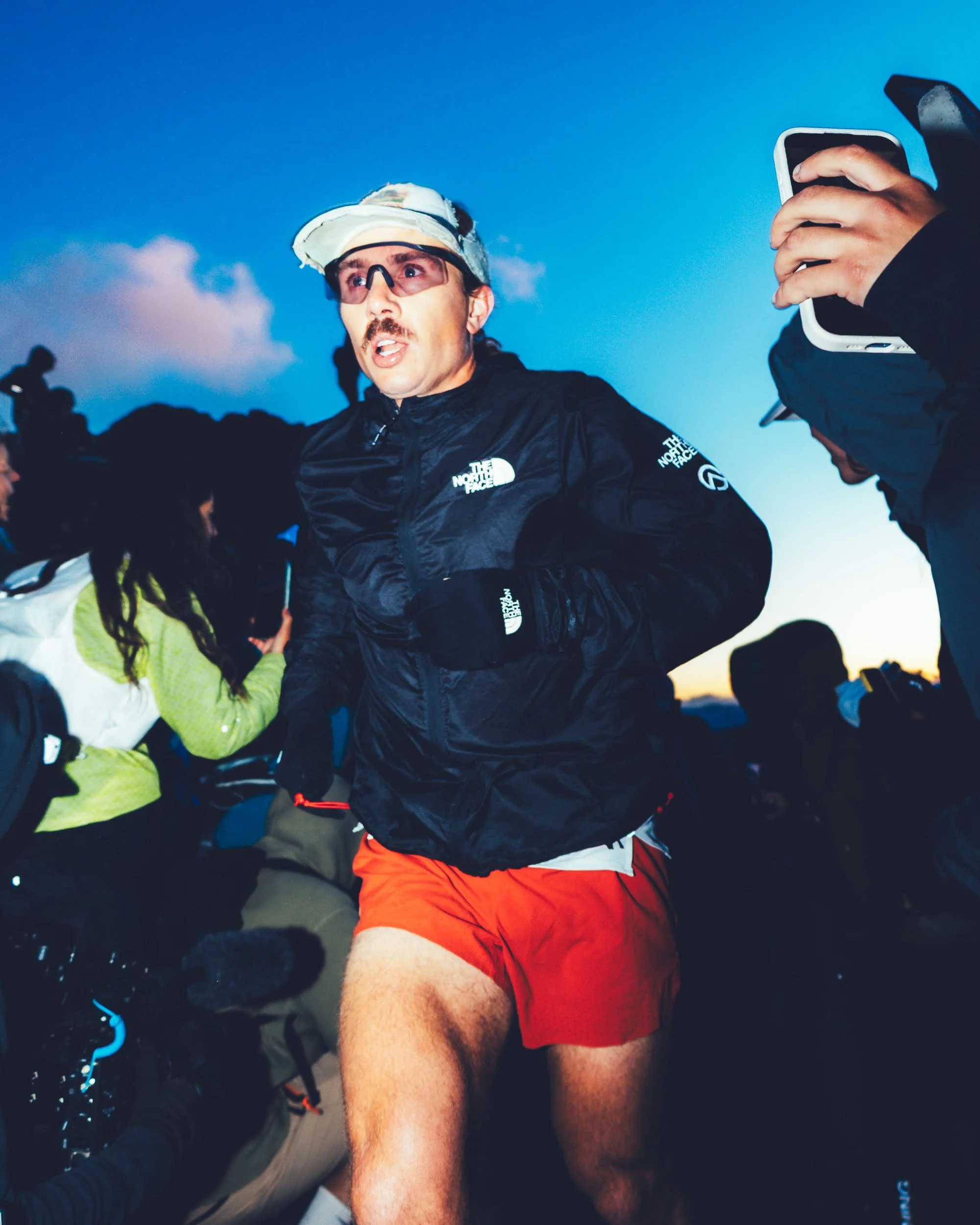 Man wearing glasses, a hat, and a black North Face jacket running outdoors at dusk, with a clear sky in the background.