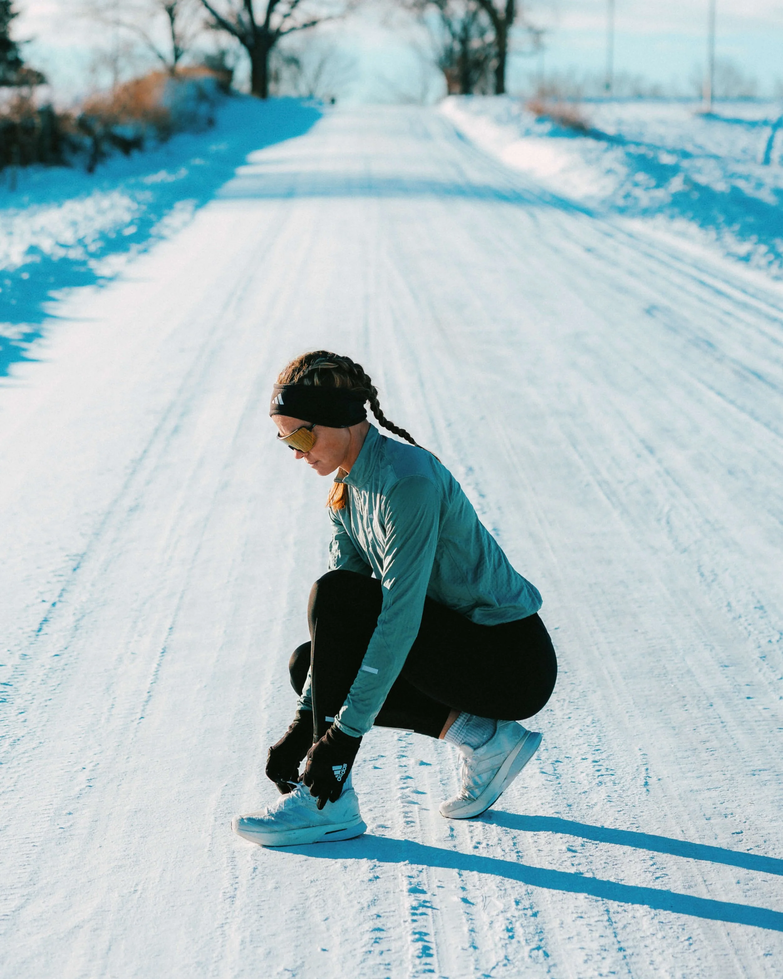 A woman in athletic clothing and white sneakers adjusting her laces on a snow-covered path in winter, with trees and a fence in the background.