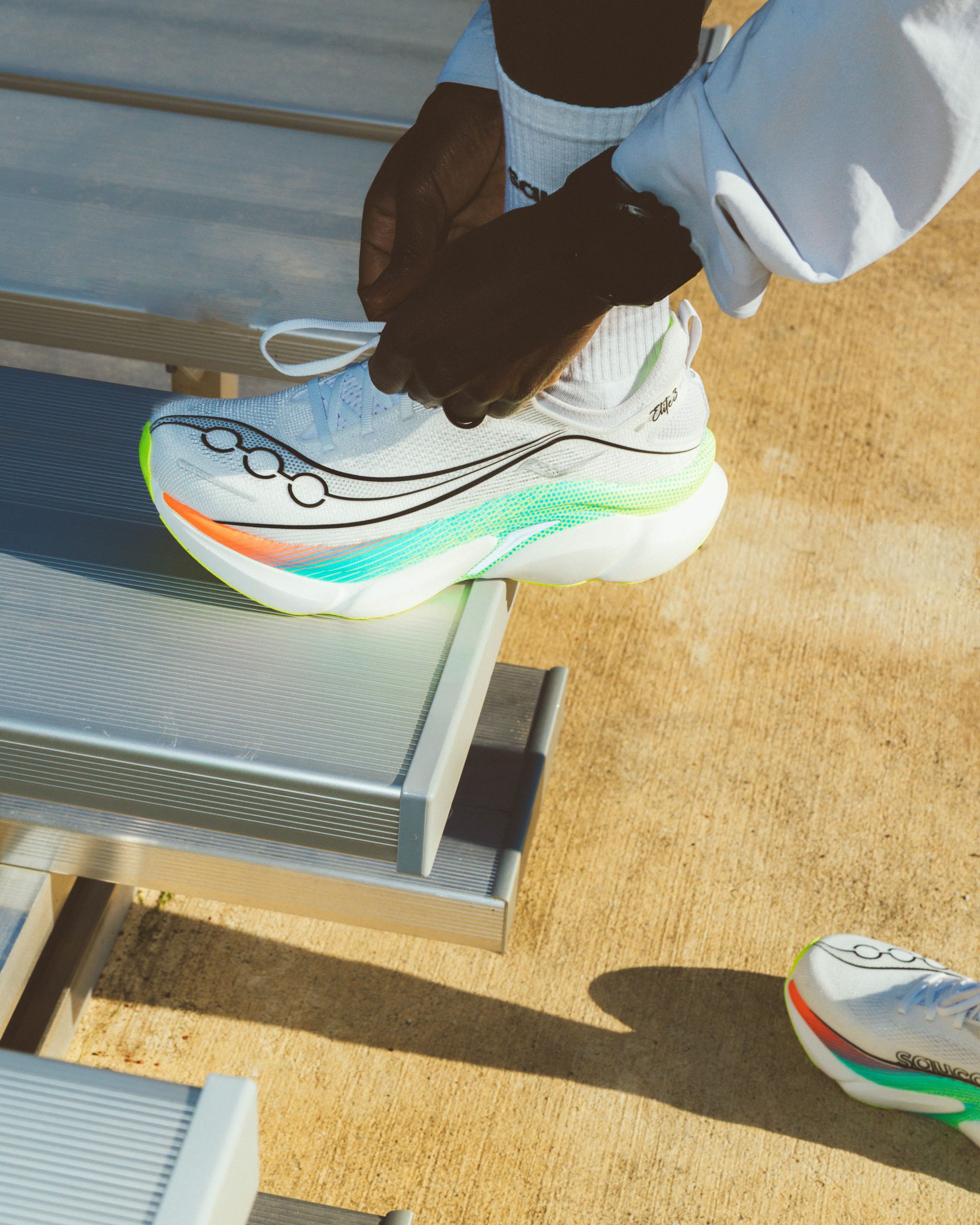 Person tying the laces of a white athletic shoe with black and colorful accents, standing outdoors on a sandy surface near a metallic structure.