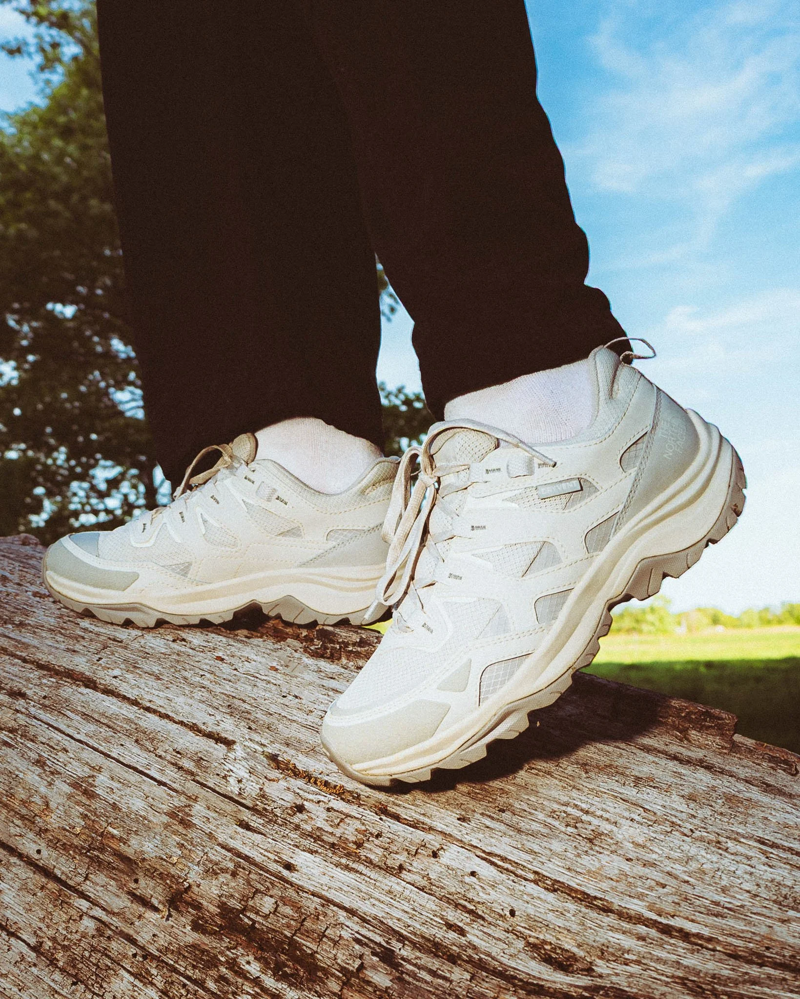 Close-up of a person's feet wearing white sneakers and black pants, standing on a weathered wooden surface outdoors with a grassy field and blue sky in the background.