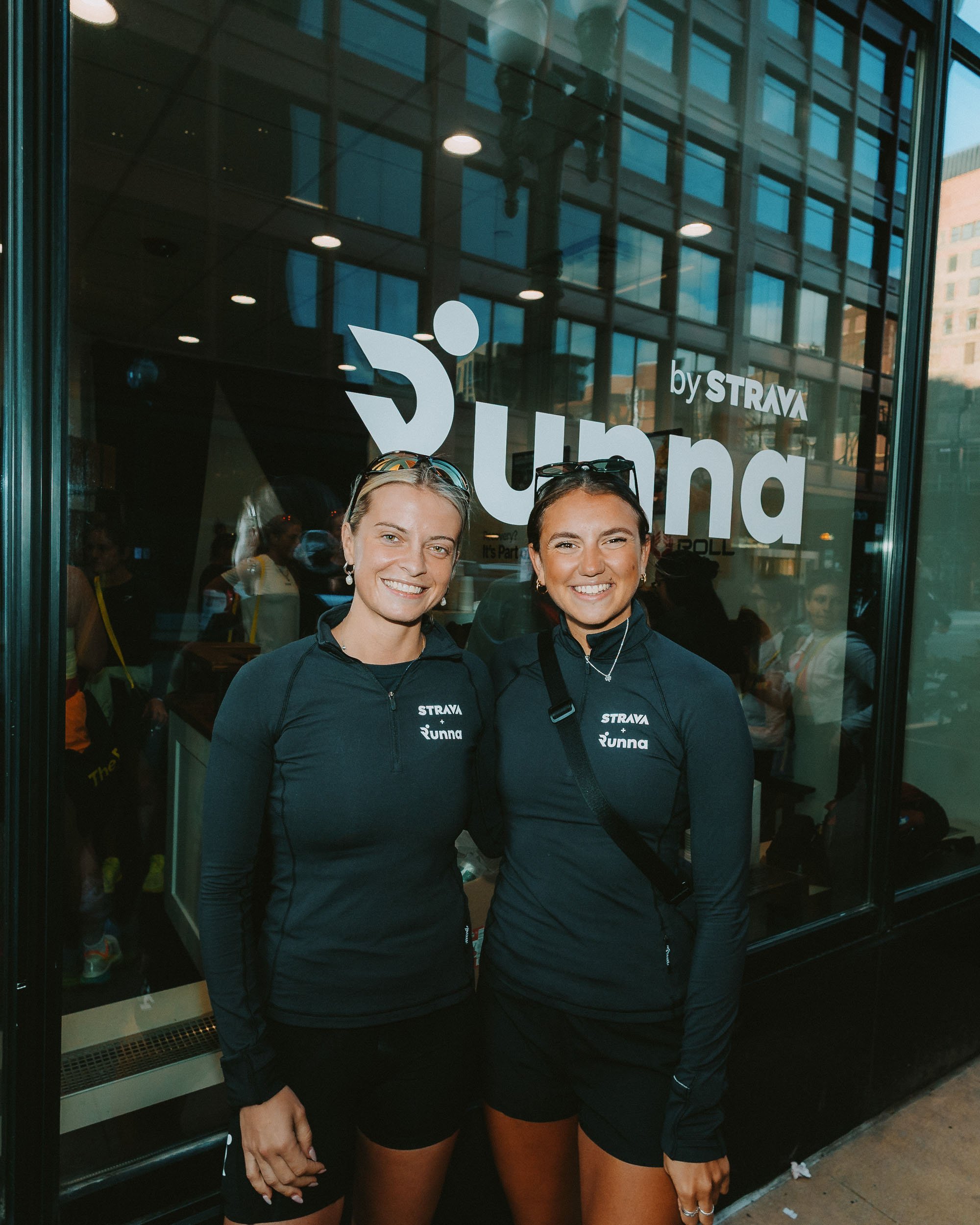 Two women wearing black jackets and shorts standing in front of a glass window with the 'by STRAVA' and 'Runna' logos, smiling at the camera.
