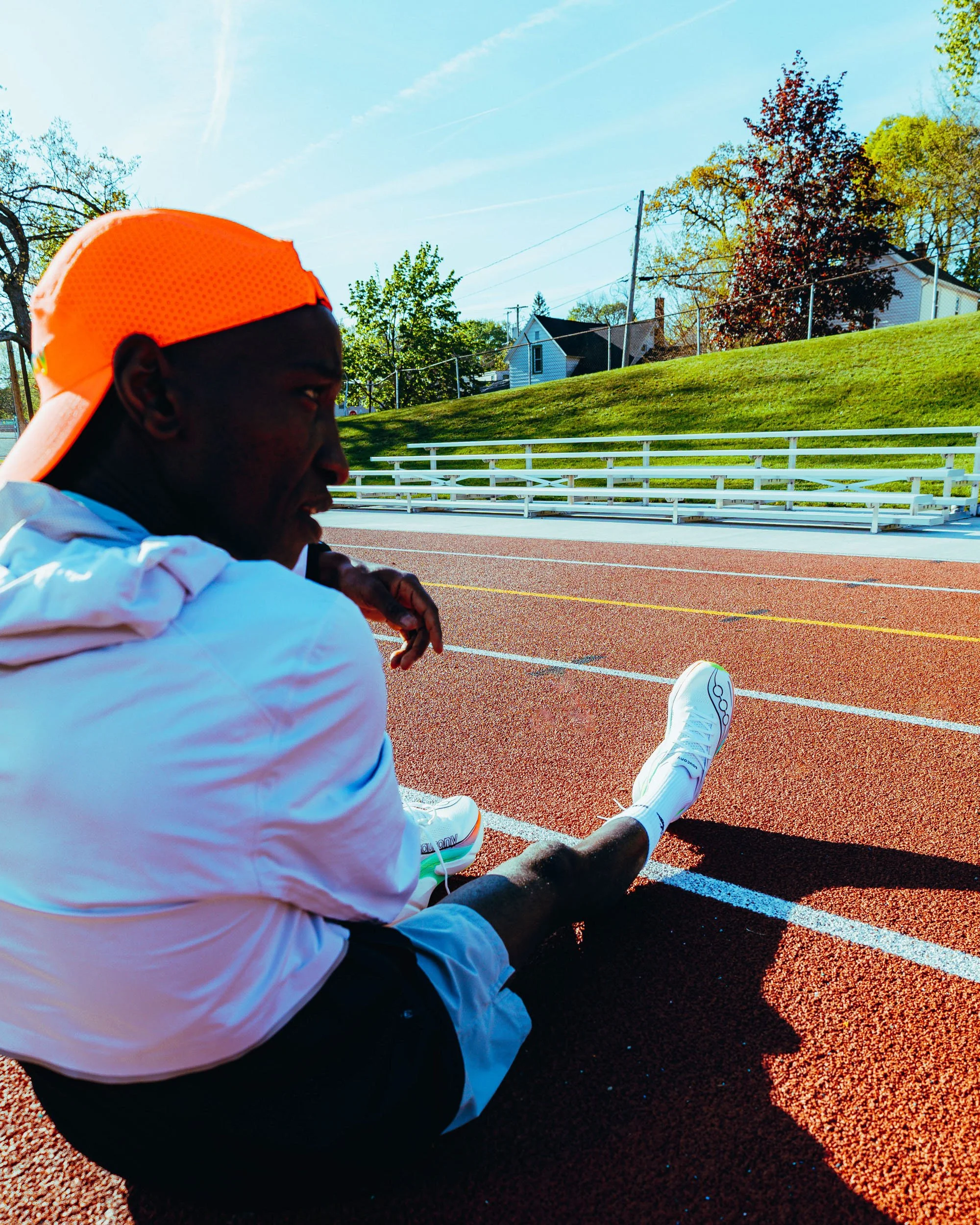 A person sitting on a running track wearing a bright orange cap, white jacket, and white athletic shoes, with trees and houses in the background on a sunny day.