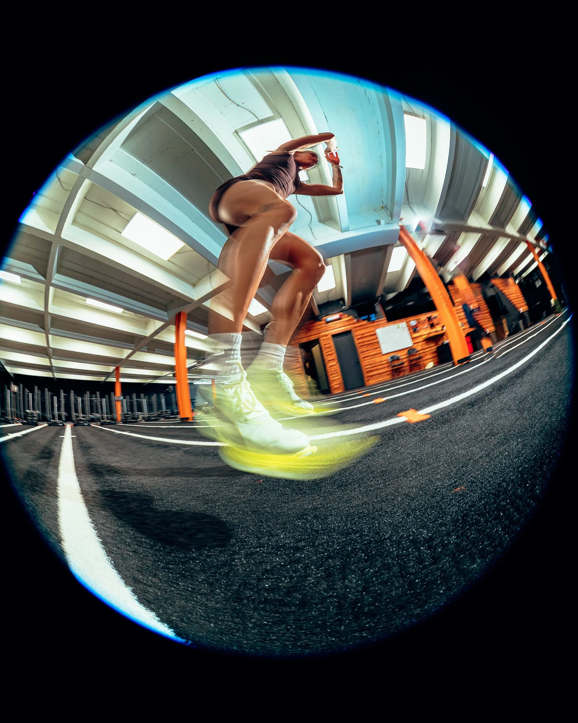 A woman in athletic clothing standing on a running track inside a gym, reflected in a shiny, circular mirror.