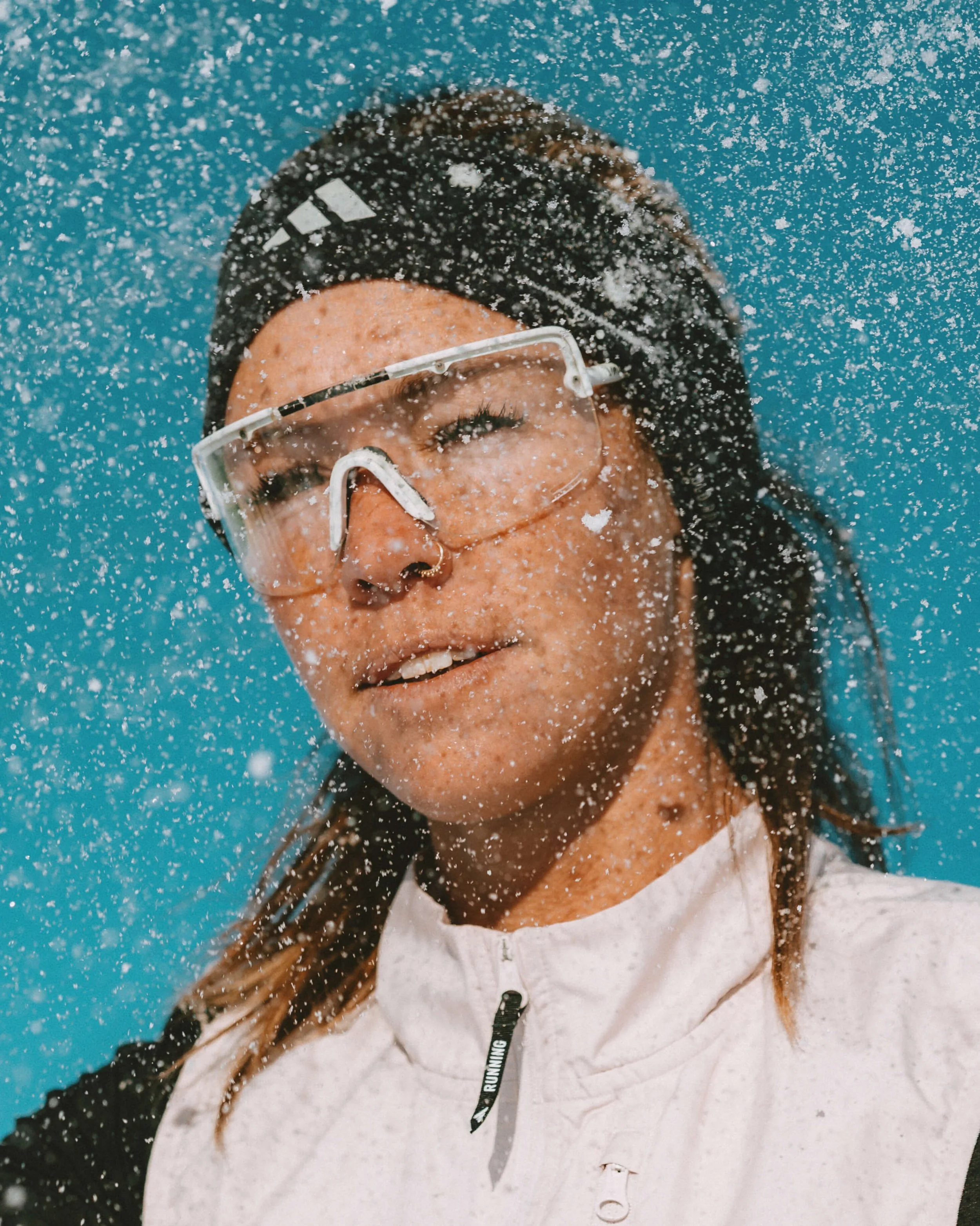 A woman with wet hair, wearing glasses and a white jacket, is submerged underwater with bubbles all around her face.