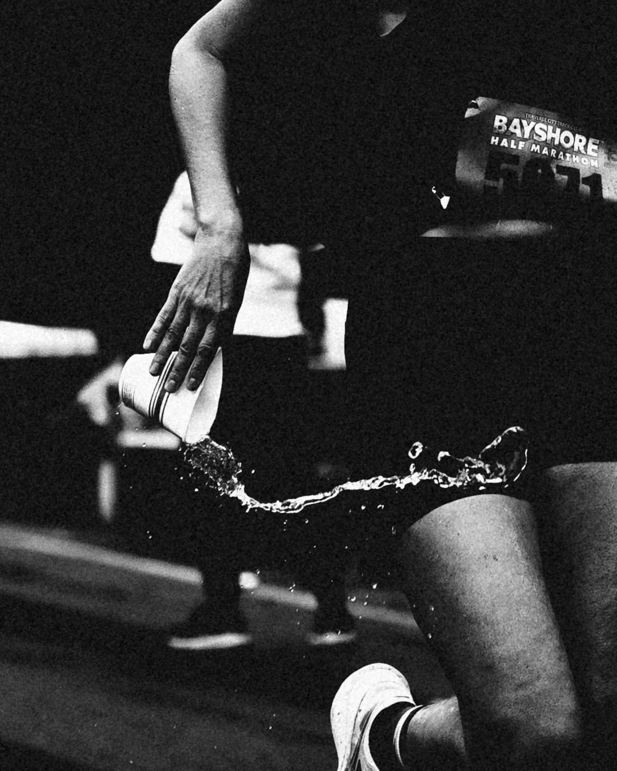 Close-up of a person's hand hitting a soccer ball on a dark surface, with part of their leg and shoe visible and a paper sign that reads "BAYSIDE HALF MARATHON" in the background.