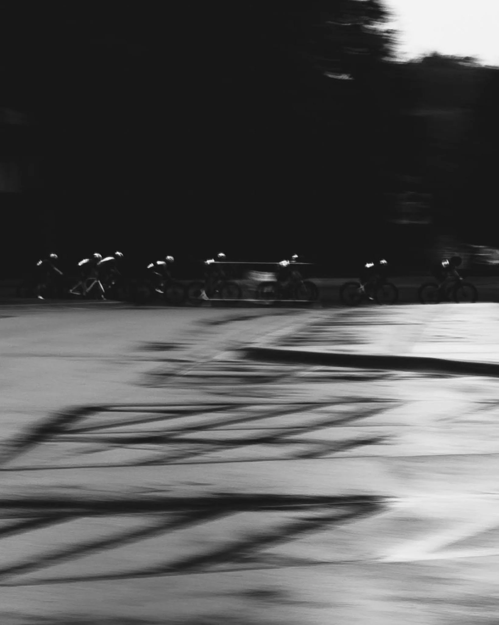 Black and white photo of a row of bicycles parked along a street at night, with blurred motion of a passing vehicle in the foreground and dark trees in the background.
