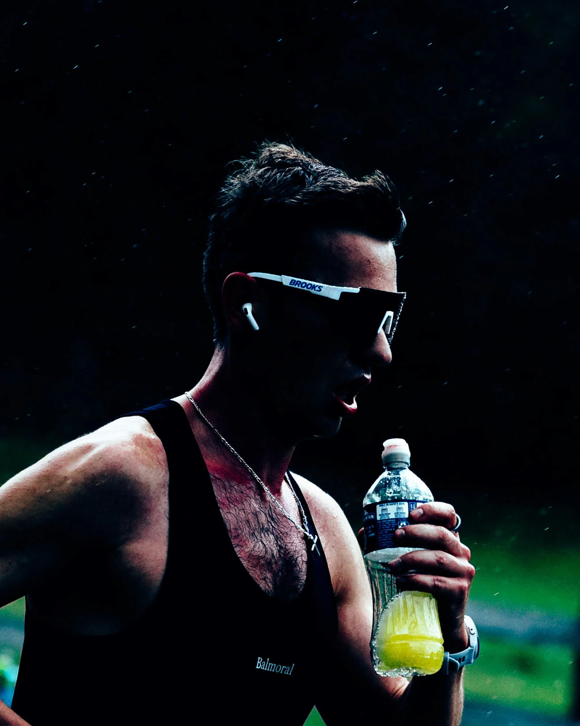 A man with dark hair and a beard, wearing sunglasses and a black tank top, is holding a bottle of water and a lemon-lime soda, running outdoors in a green park during the daytime.
