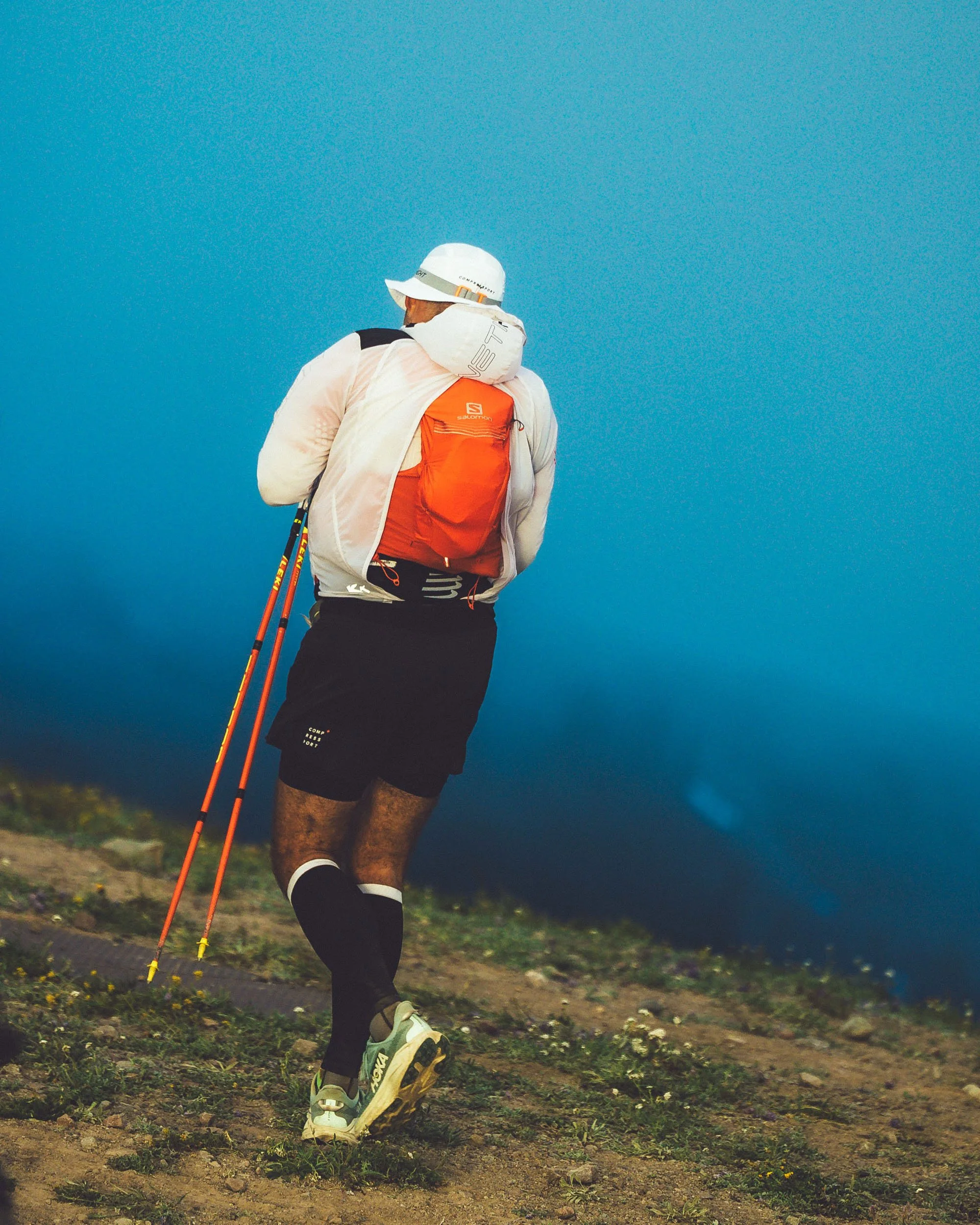 A man running outdoors on a trail, wearing a white hat, a white jacket, dark shorts, and running shoes, carrying a backpack and using trekking poles against a blue sky.