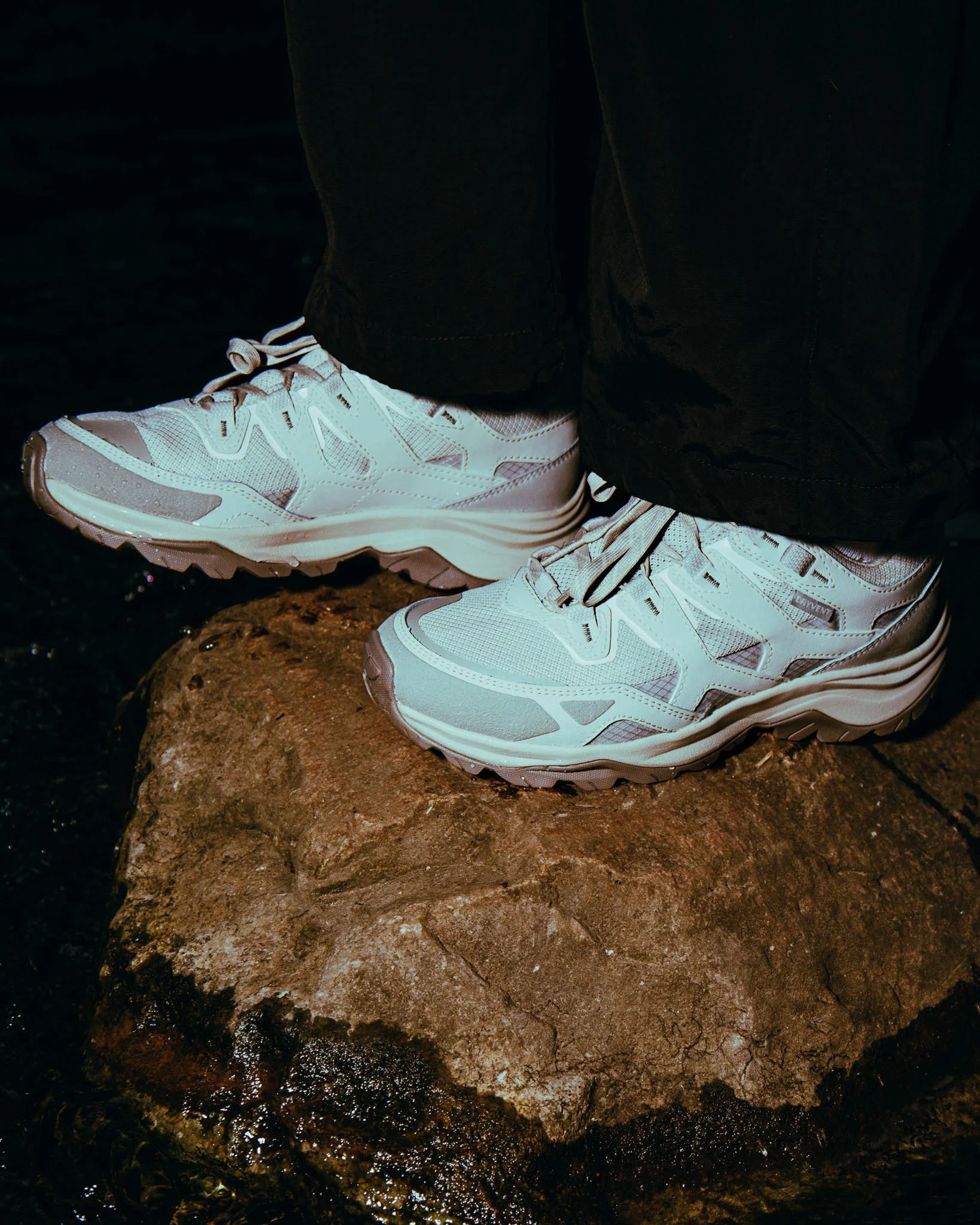 Close-up of a person's legs into black pants and white sneakers standing on a large, uneven, rocky surface with water droplets. The rocky surface appears damp.