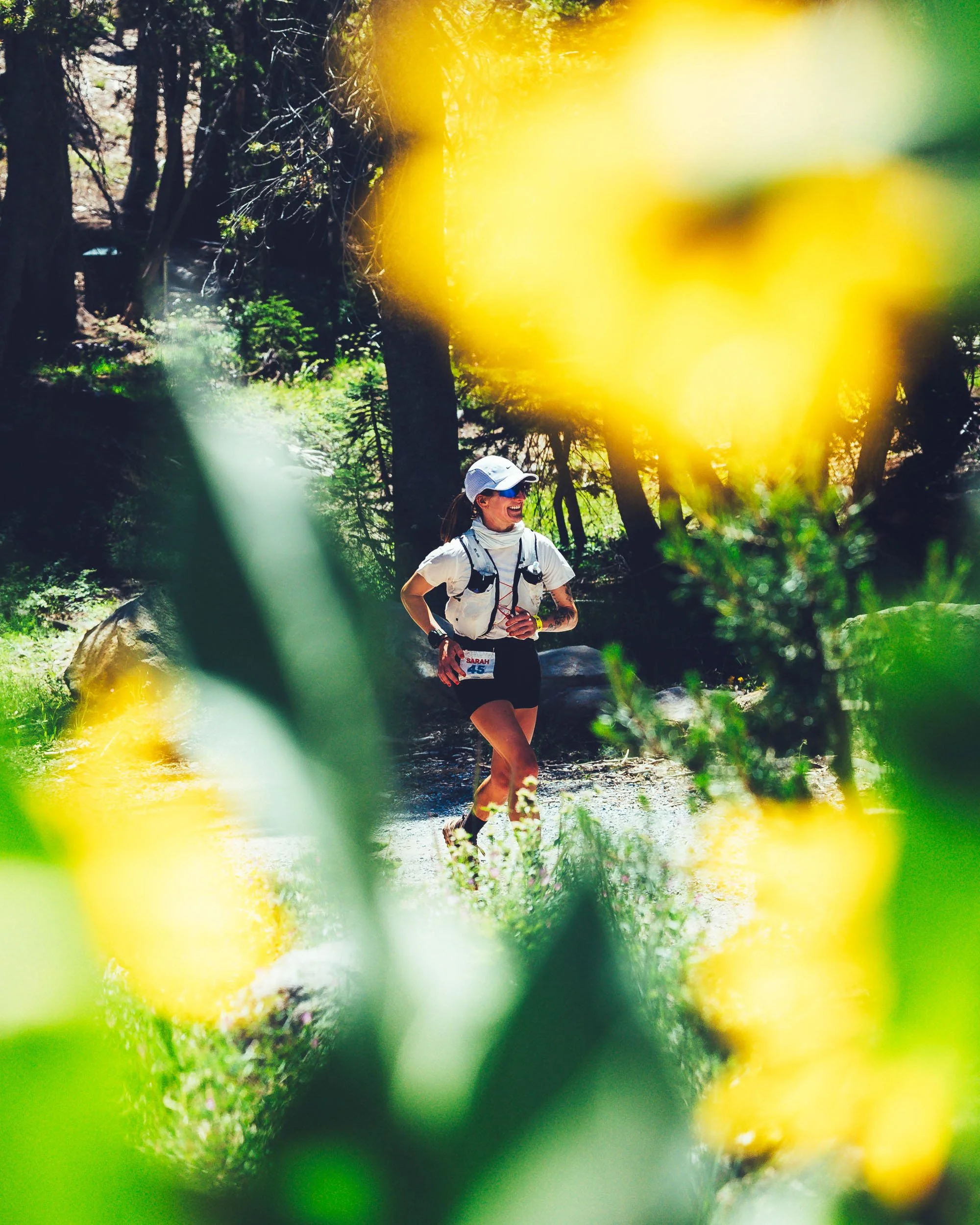 A woman with a white cap and a white hiking shirt, smiling as she runs on a trail in a forested area during the day. She is wearing a numbered bib, indicating she is participating in a race or marathon.