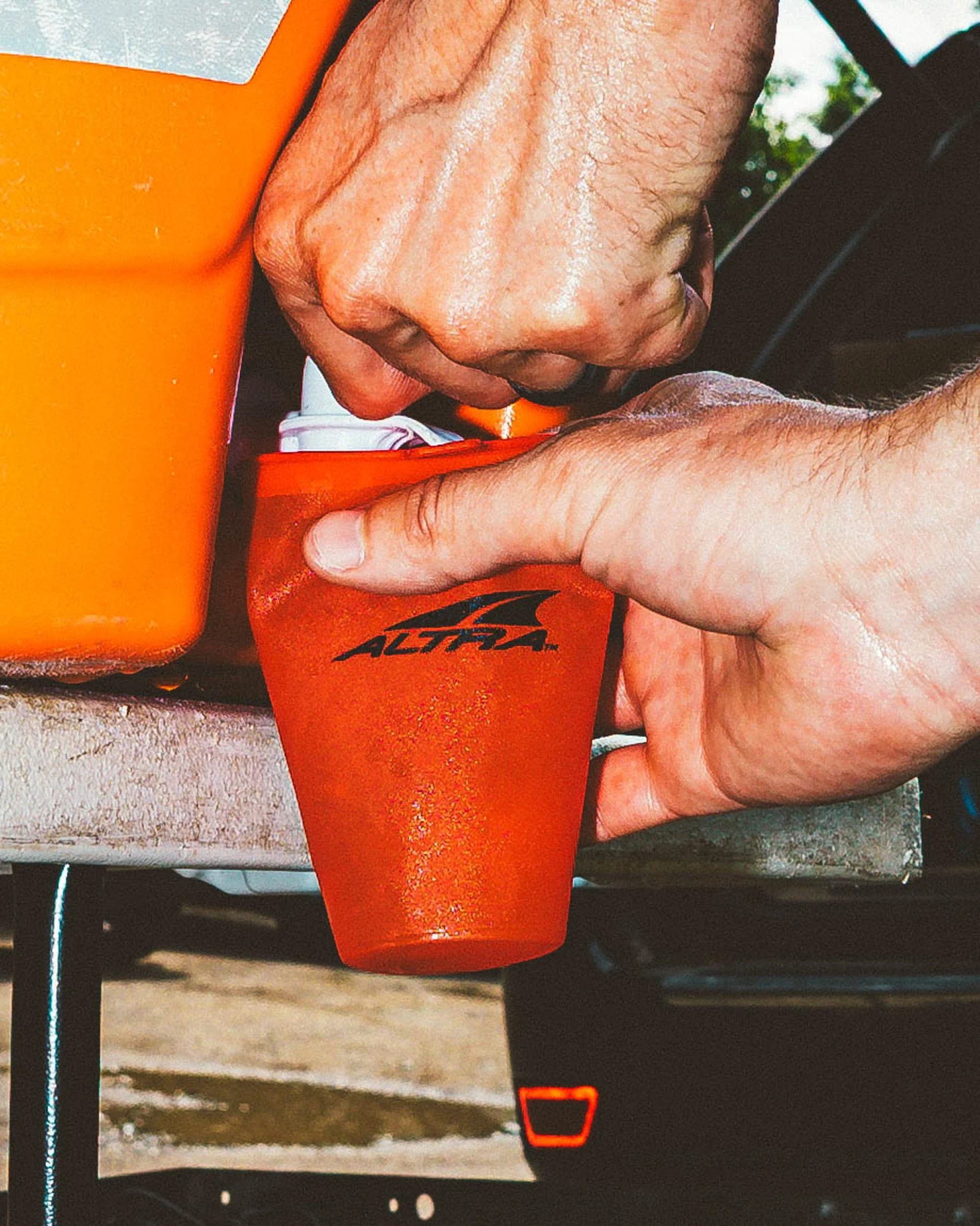 A person is filling up a bottle with orange liquid from a larger container, with a background of an outdoor setting.