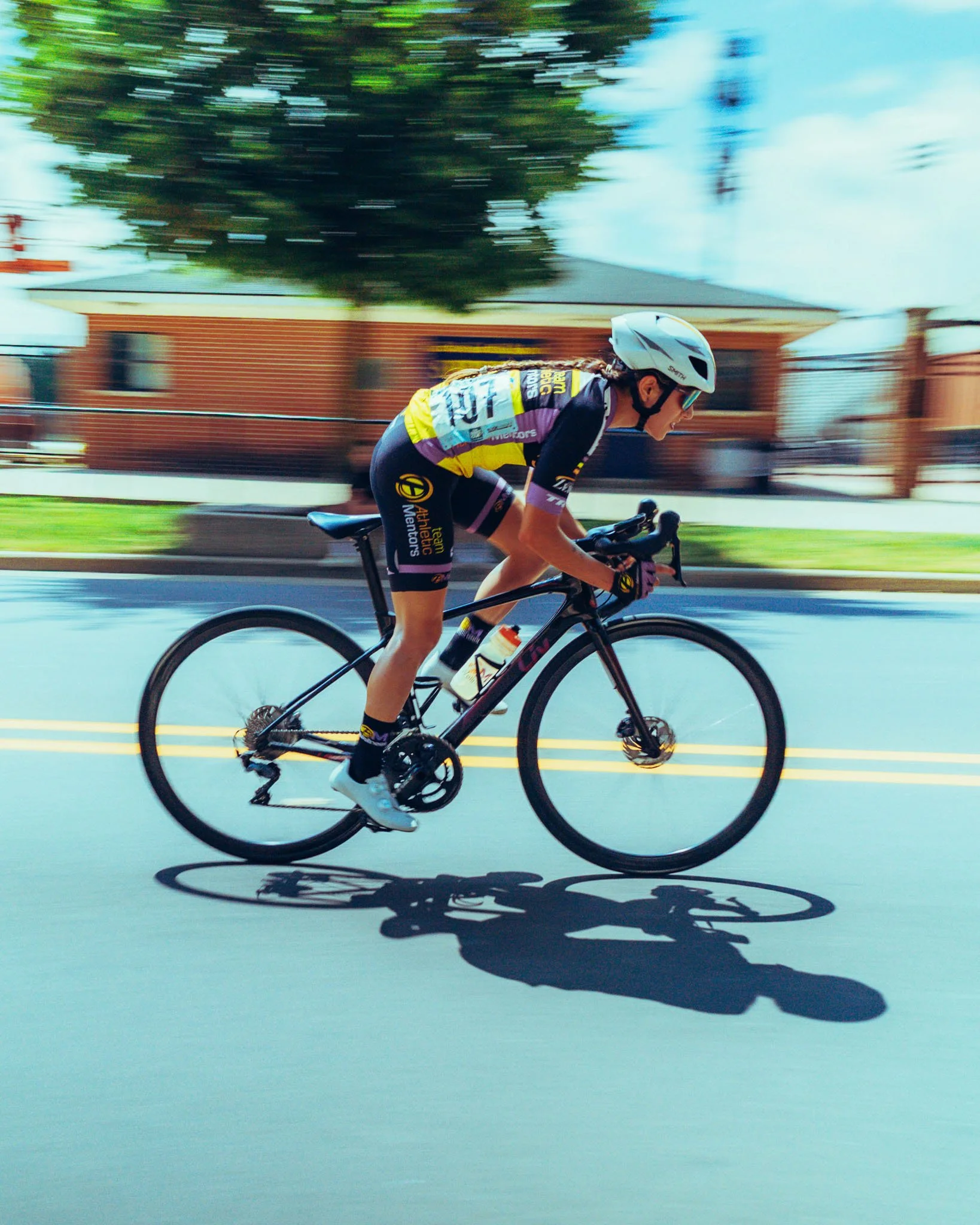 A female cyclist wearing a helmet and colorful sportswear riding a black road bike on a street with blurred background of trees and houses, casting a shadow on the pavement.