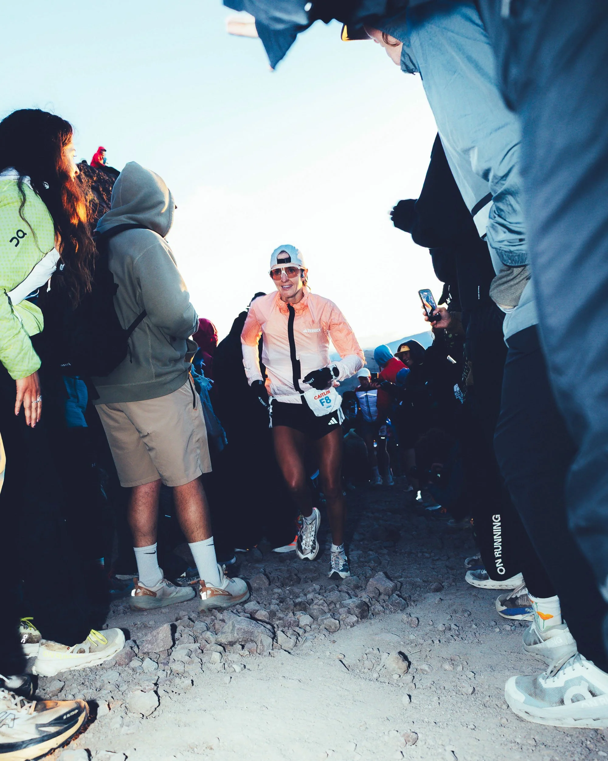 Group of runners standing on rocky mountain trail, some bending forward, dressed in athletic gear, preparing for race or training, with a woman in the center wearing a white cap, pink jacket and sunglasses.