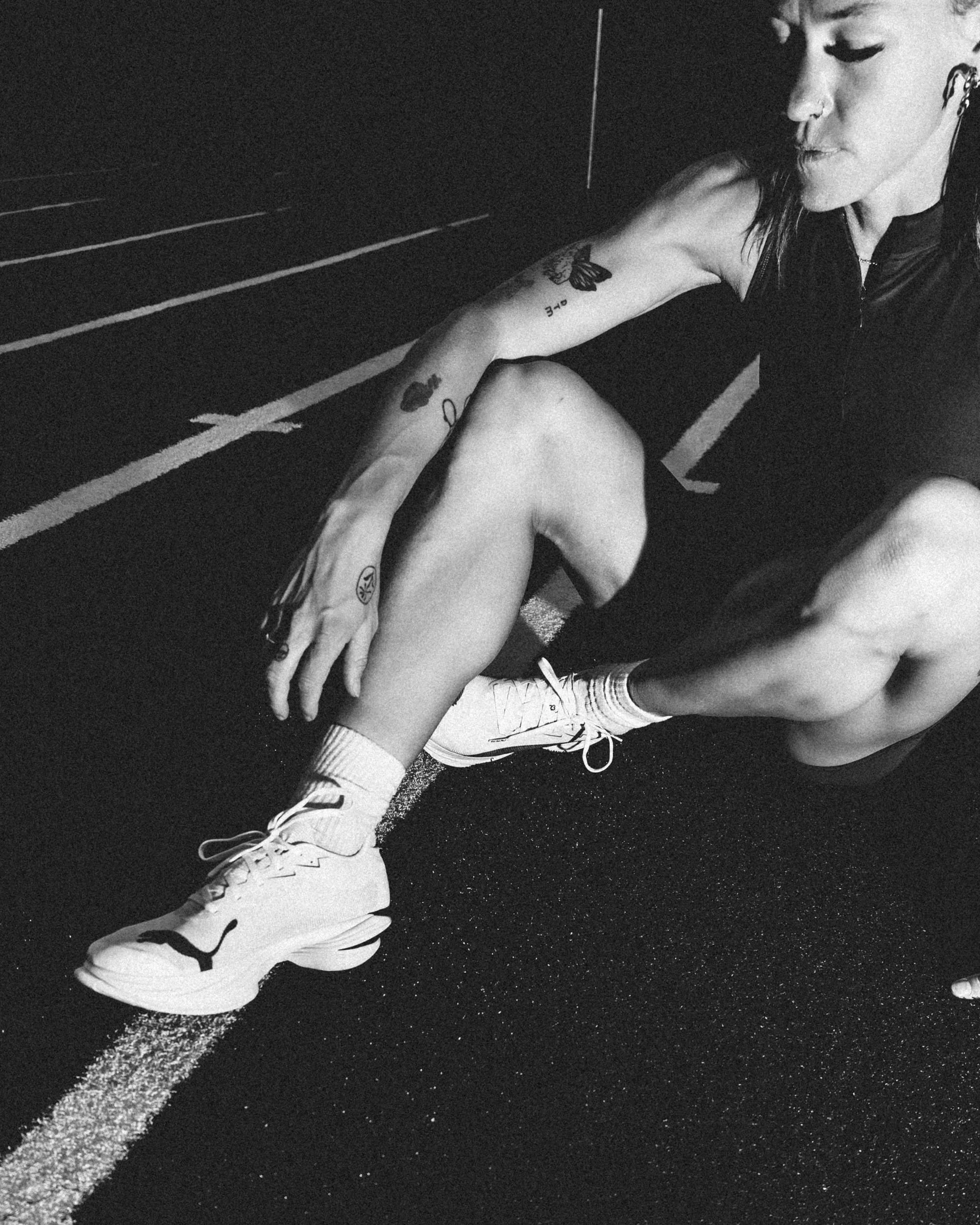 A person with tattoos on their arm and wearing athletic shoes sits on a running track, looking down.