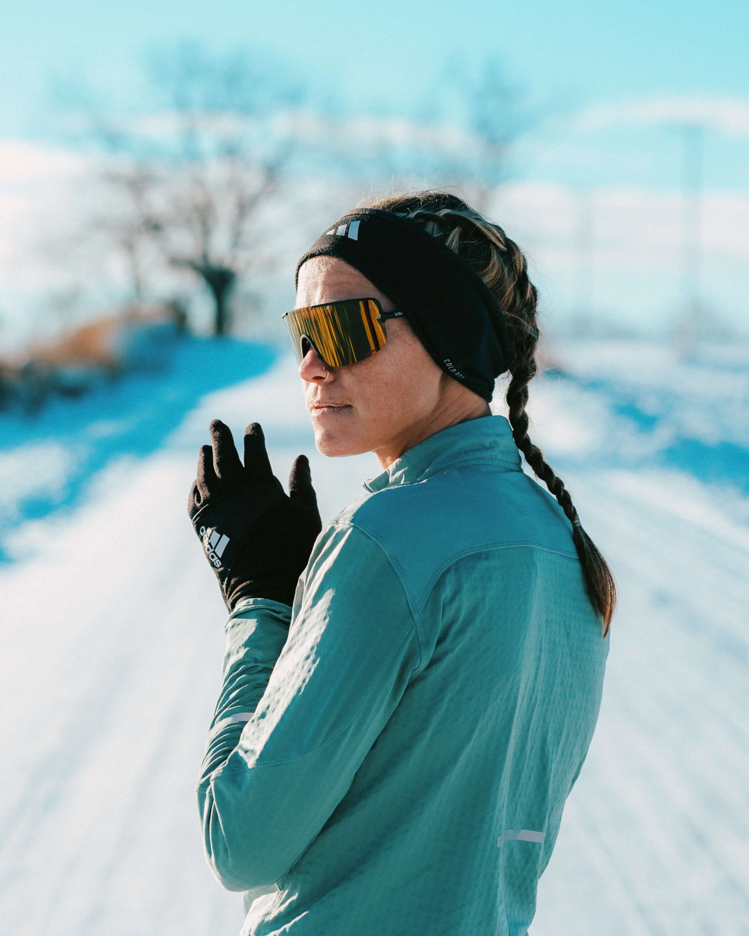 Close-up of a woman in winter sports gear outdoors, wearing reflective sports sunglasses, a headband, and a jacket, with an icy landscape and snow-covered trees in the background.