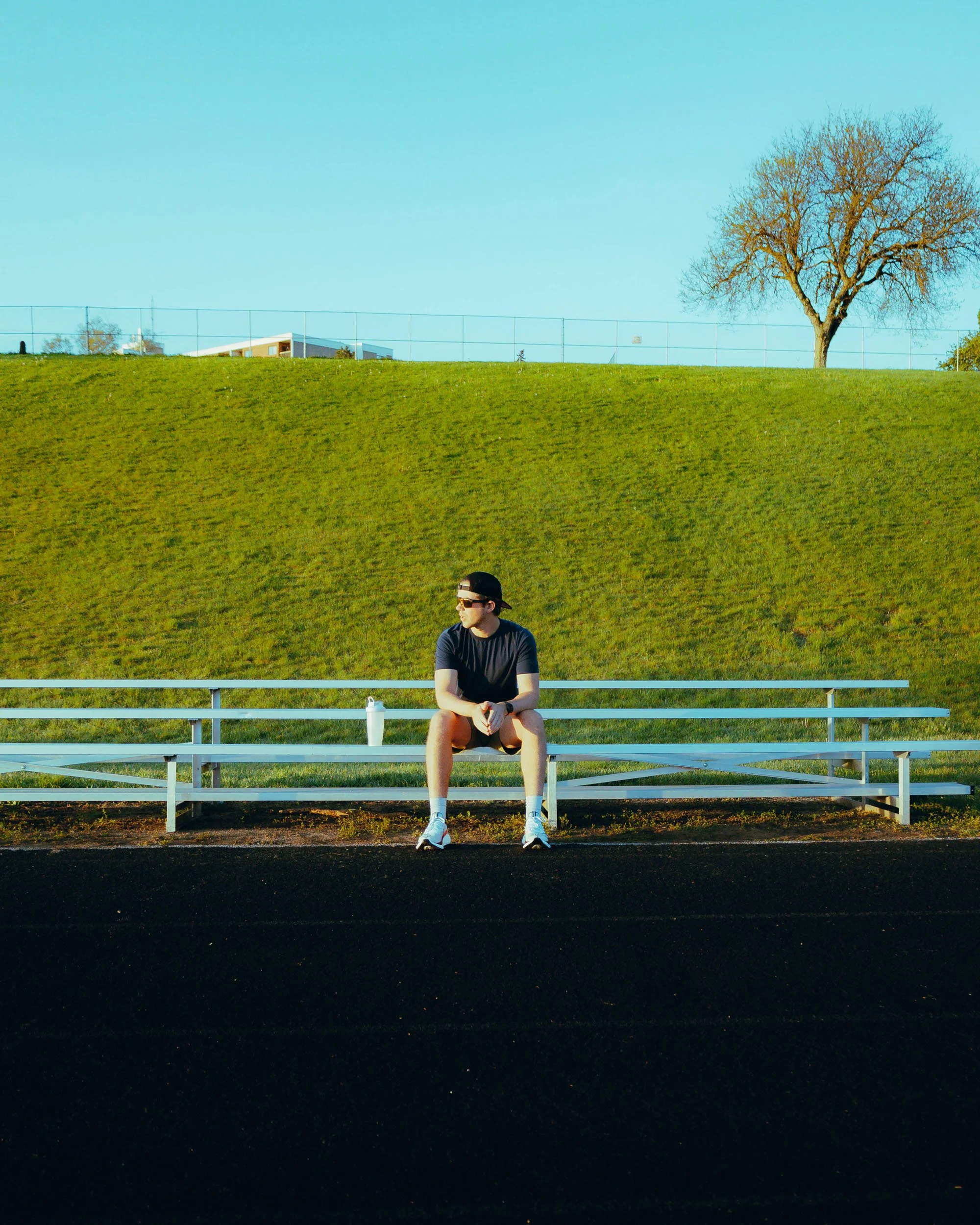 A young man sitting on a white bleacher in front of a grassy hill with a single tree and a building in the background; the sky is clear and blue.