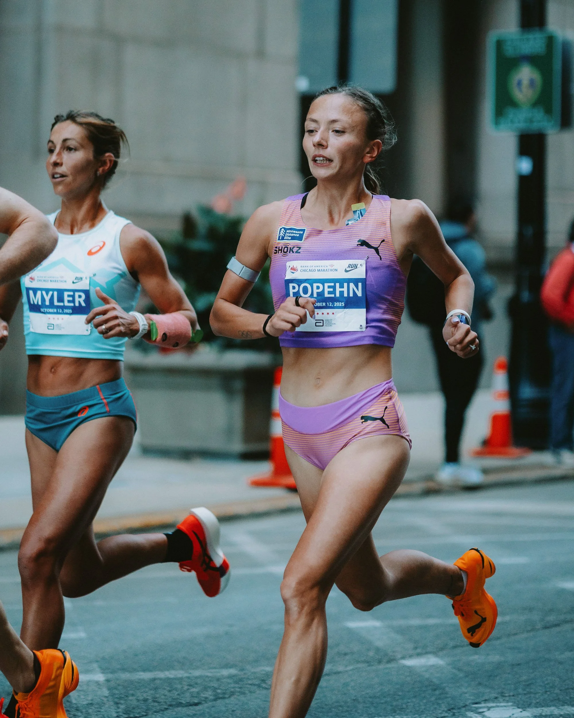 Two marathon runners, one woman wearing a purple sports bra and shorts, and the other woman wearing a white tank top and blue shorts, running on a city street during a race.