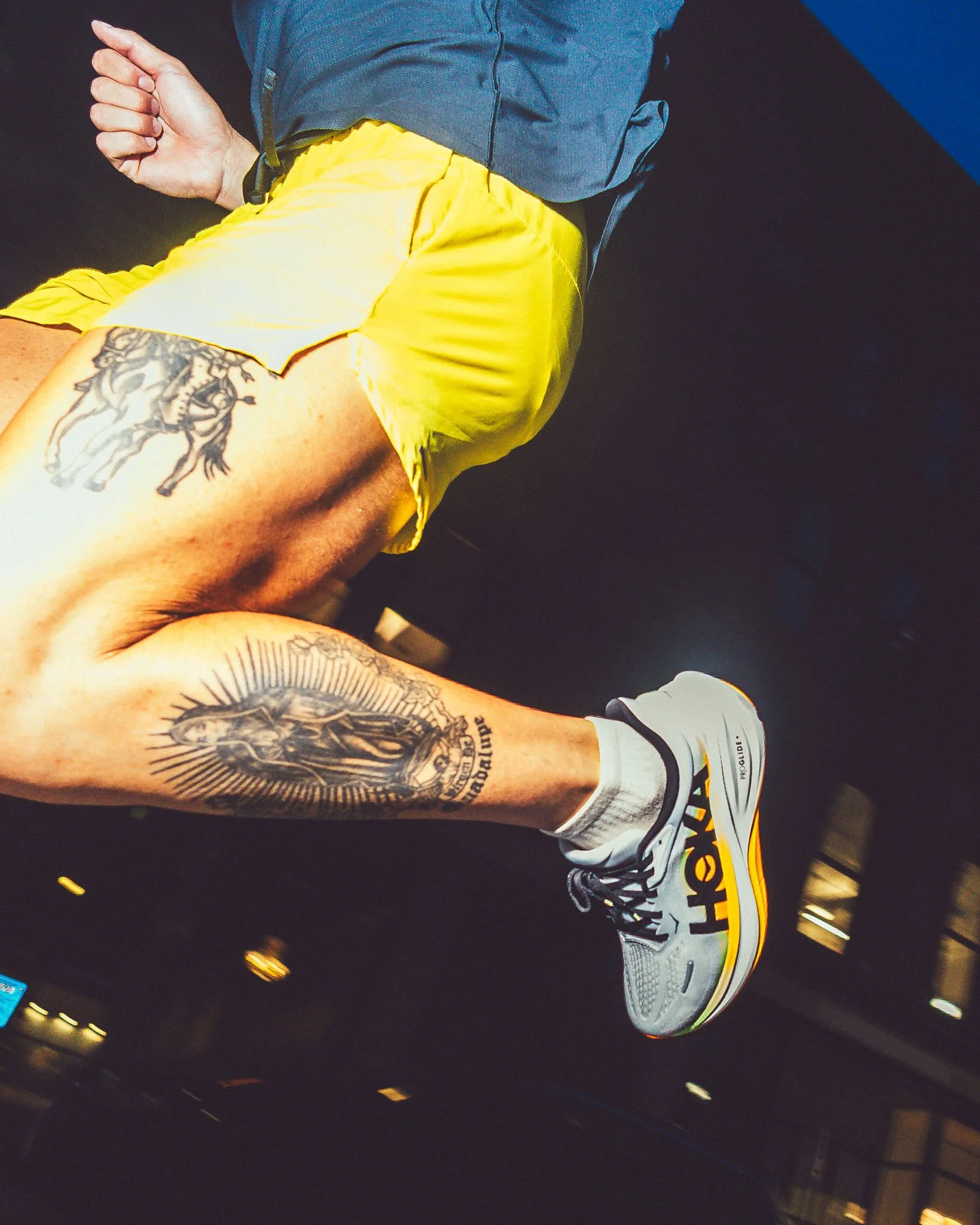 Close-up of a person wearing a gray shirt, bright yellow shorts, white sneakers with yellow and black accents, and white socks. The person's tattooed arm is visible, and they are holding their thumb out with a thumbs-up gesture.