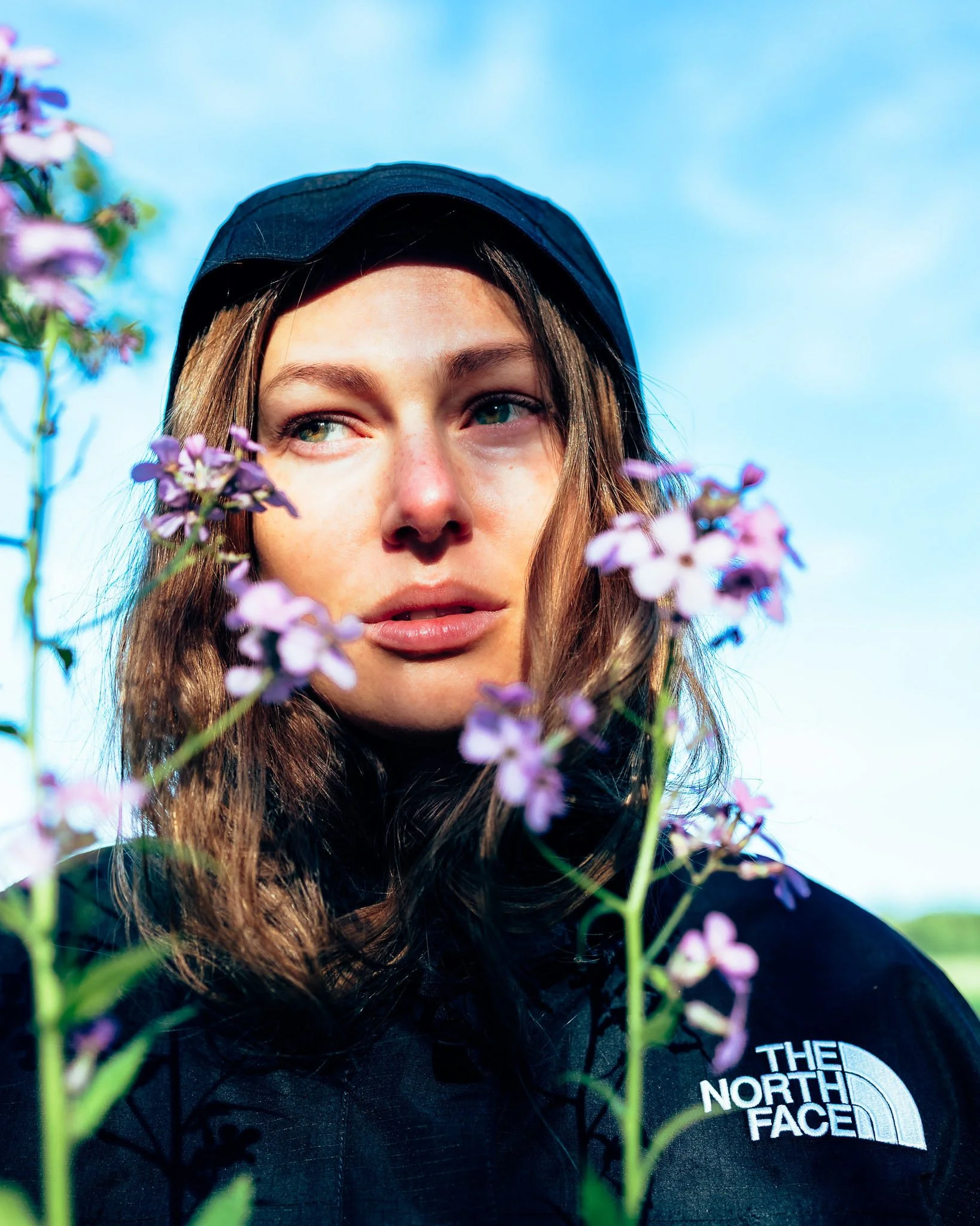 Close-up of a young woman with brown hair wearing a black cap and a black The North Face jacket, standing among pink flowers against a clear blue sky.