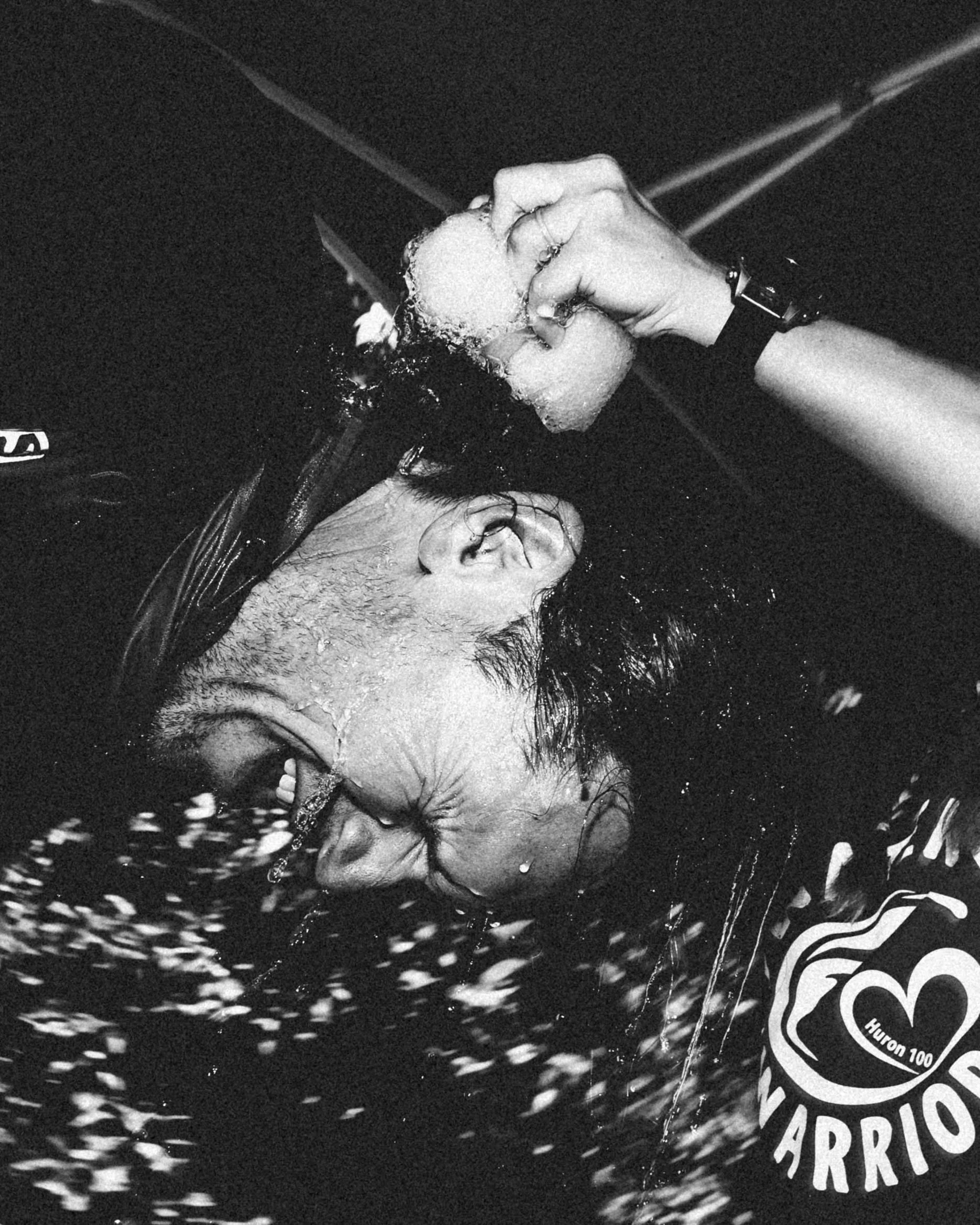 A person is holding an ice cube in one hand, with water splashing around them. The person is wearing glasses, have wet hair, and are wearing a dark shirt and a wristwatch. The background is dark and appears to be in a setting related to water or cold