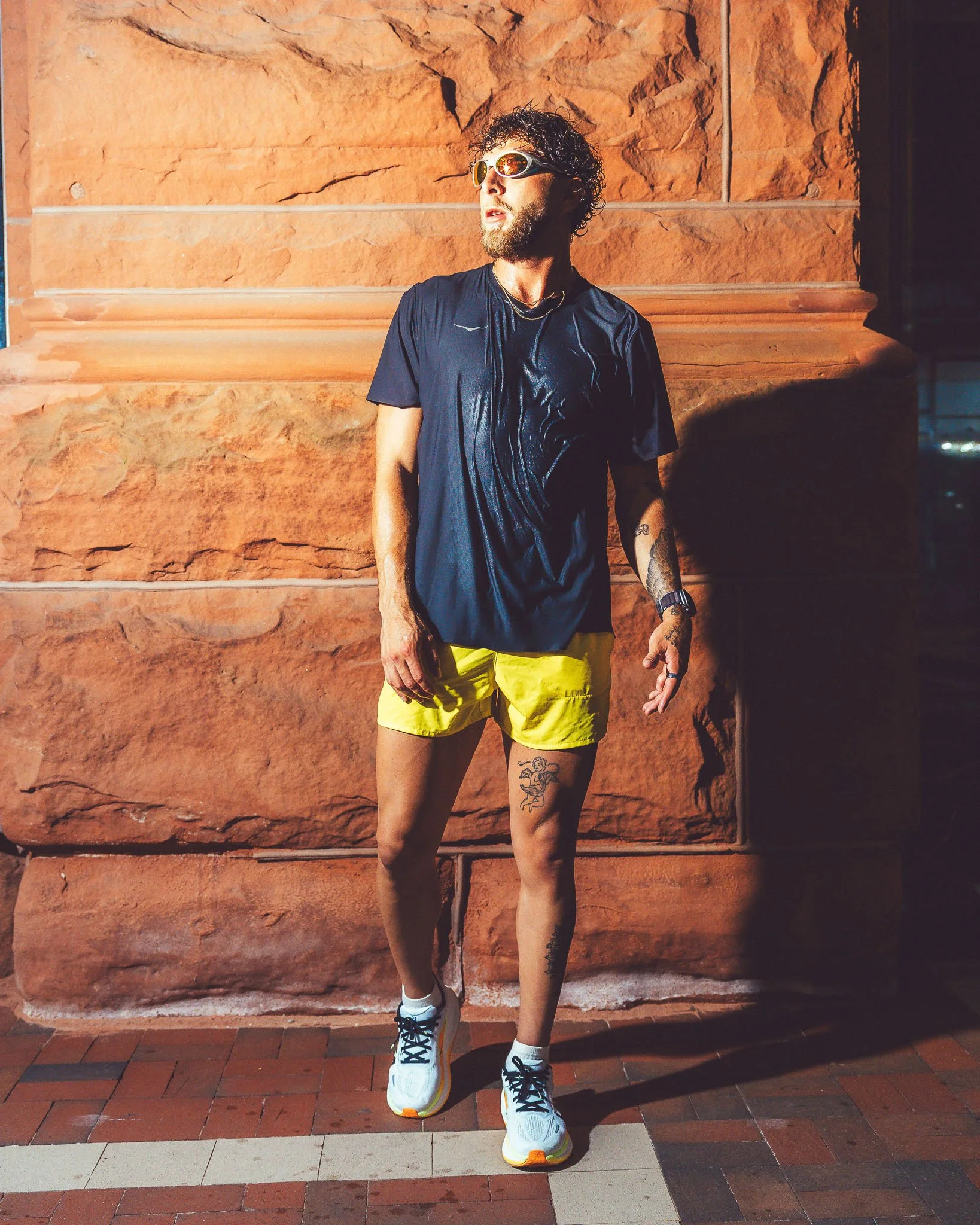 A man standing in front of a red brick wall, wearing sunglasses, a black t-shirt, yellow shorts, white sneakers, and displaying tattoos on his legs and arm.