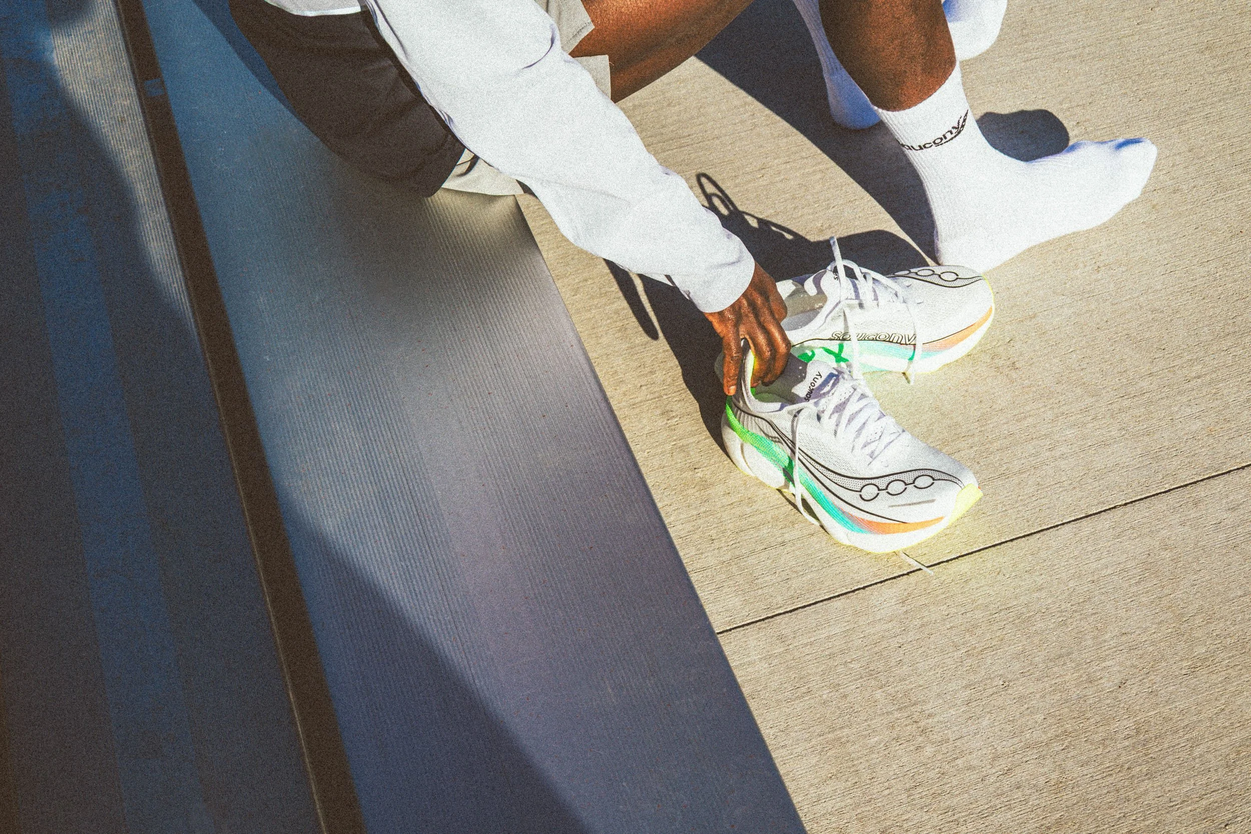 A person wearing white athletic shoes with colorful accents, white socks, white and black athletic clothing, and brown shorts, sitting on a bench on a wooden outdoor surface.
