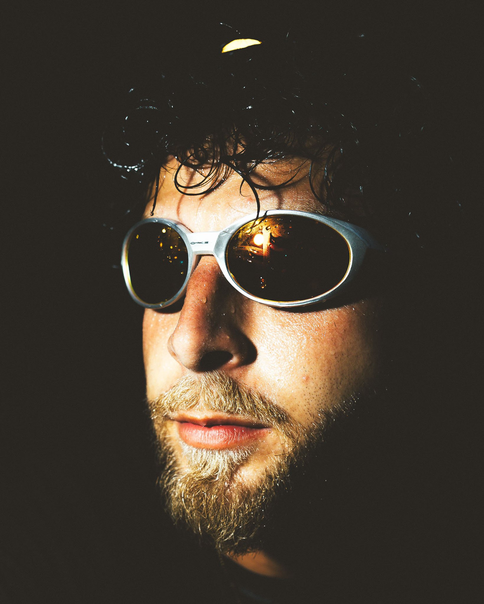 A close-up of a person's face with dark, wet hair, wearing round sunglasses reflecting a sunset scene, and a beard. The background is dark.