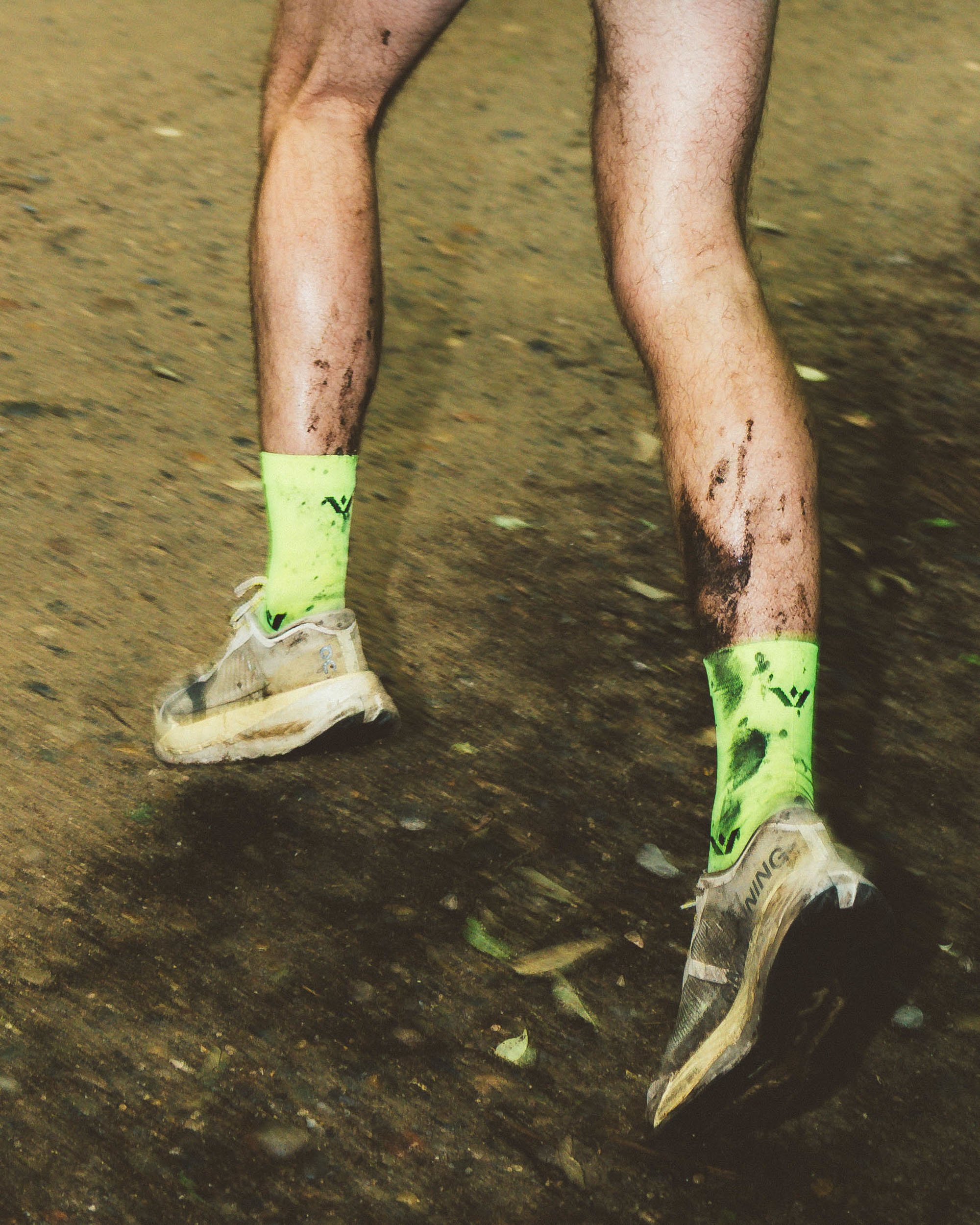 Close-up of a runner's muddy legs and shoes on a dirt trail, wearing triathlon gear including bright yellow compression socks and white running shoes.