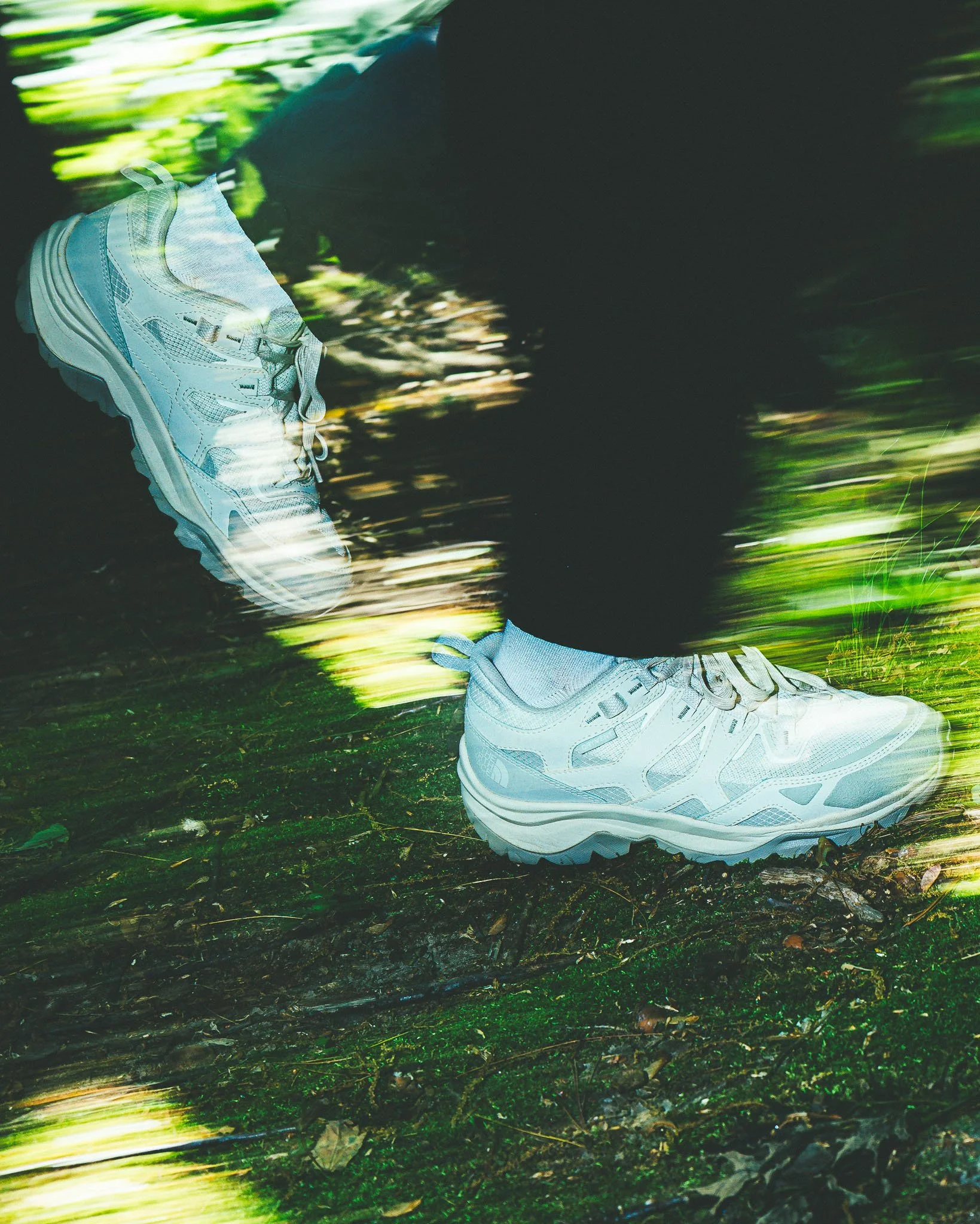 A person running outdoors wearing white athletic shoes, dark pants, and a black shirt, with a blurred background of green trees and grass.