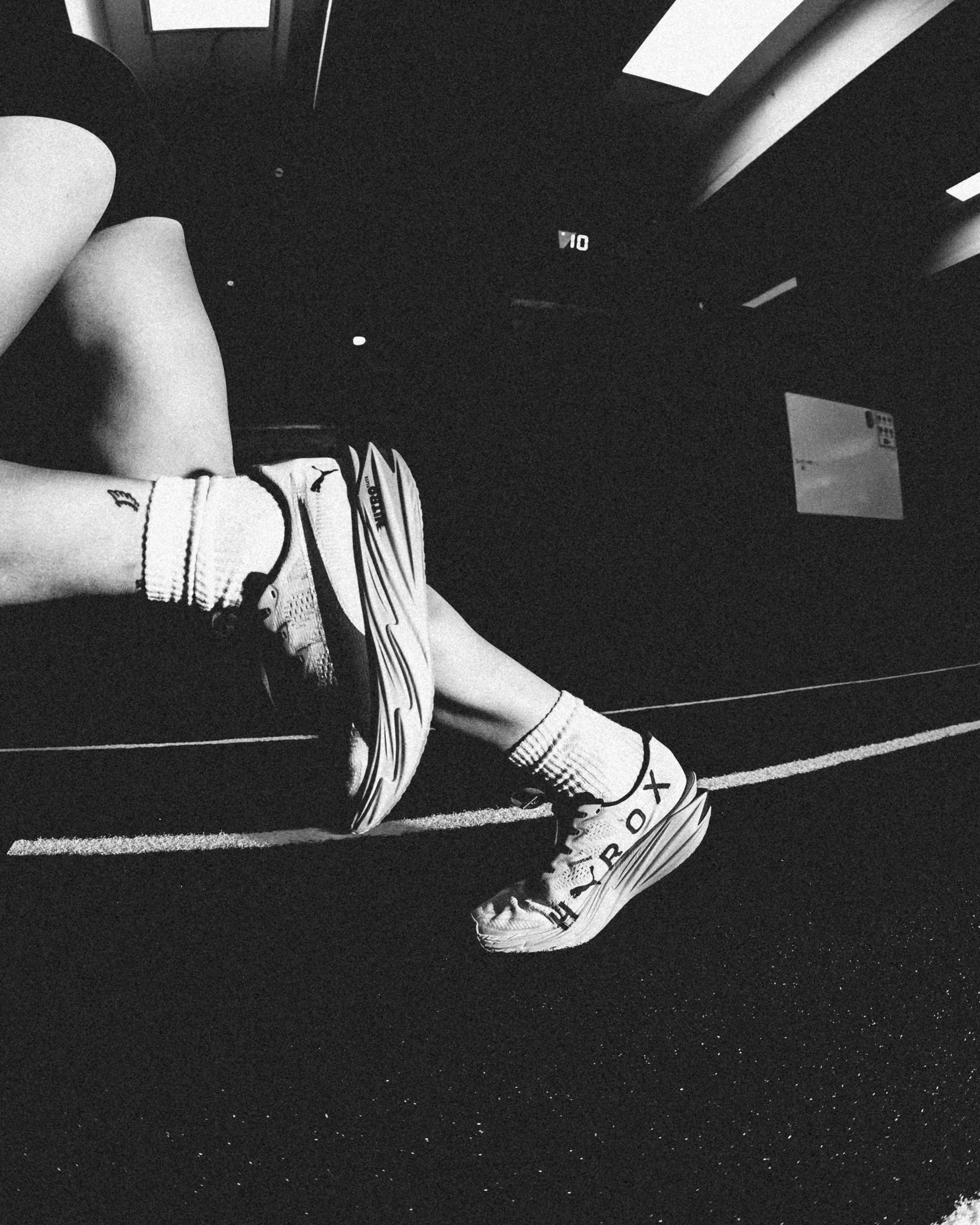 A person sitting on a black gym floor, wearing athletic clothing, including sneakers and socks, with their legs extended. The floor has white speckles and lines, and there is a whiteboard or panel on the wall in the background.
