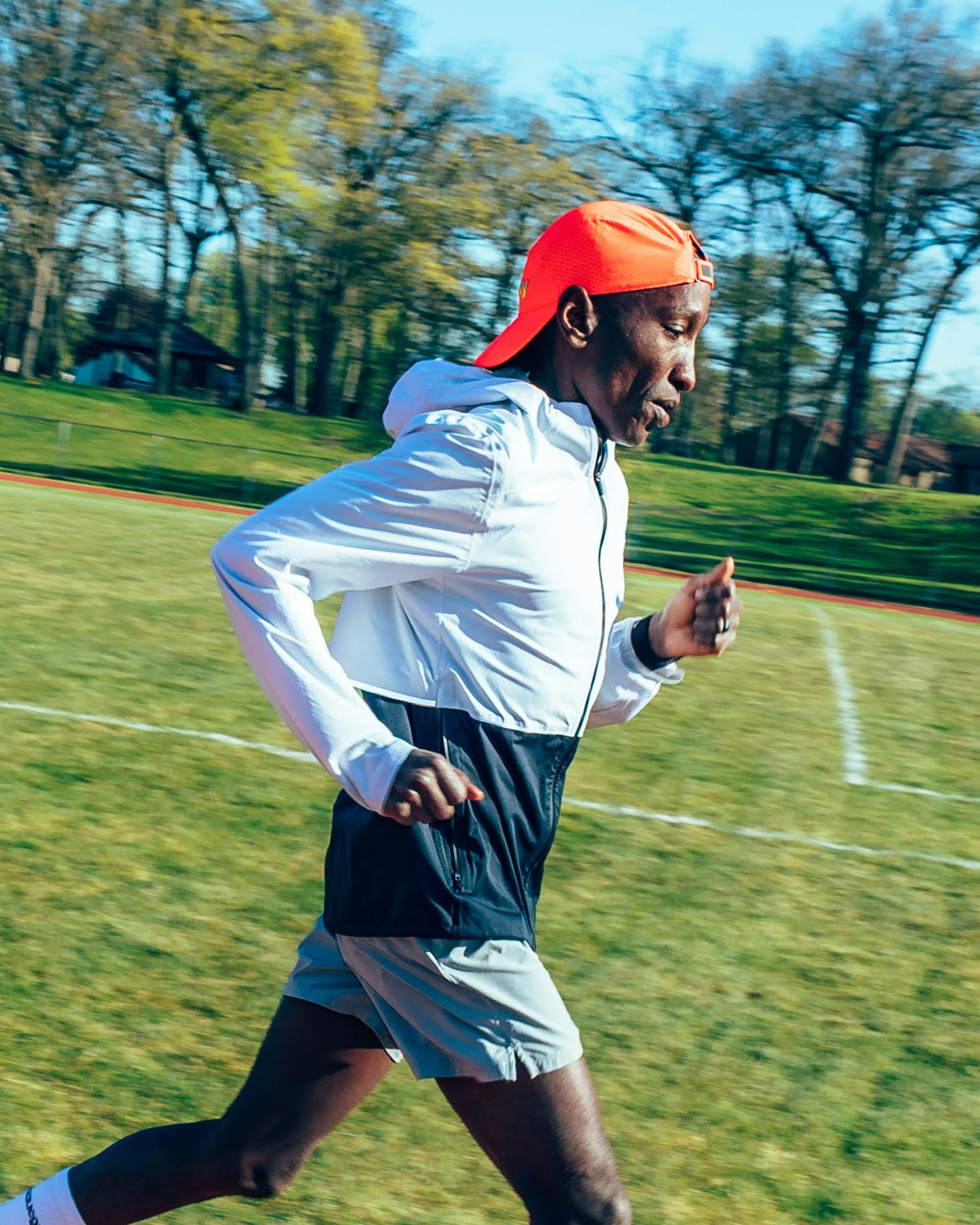 A man running outdoors on a grassy field with trees and houses in the background, wearing a red cap, white jacket, and black shorts.