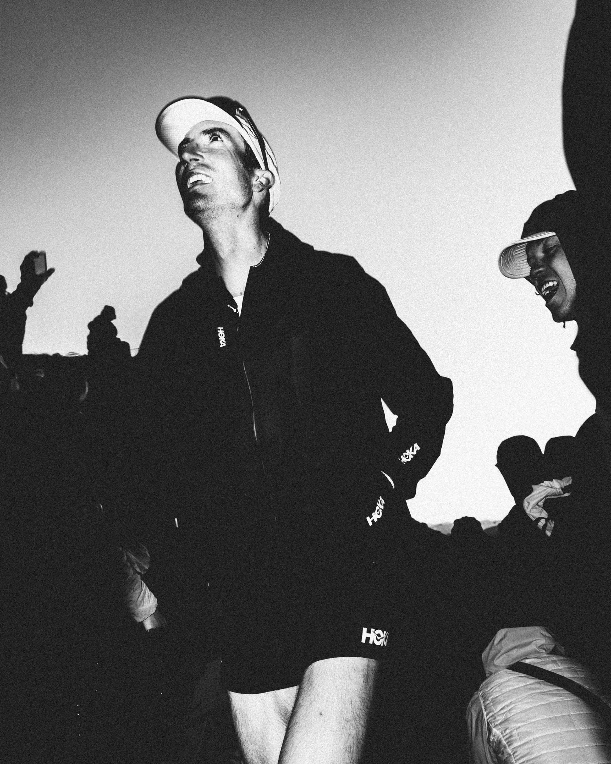 Black and white photo of two young men on a mountain or hilltop, one wearing a cap and laughing, the other wearing a hoodie and cap, sitting with their legs extended.