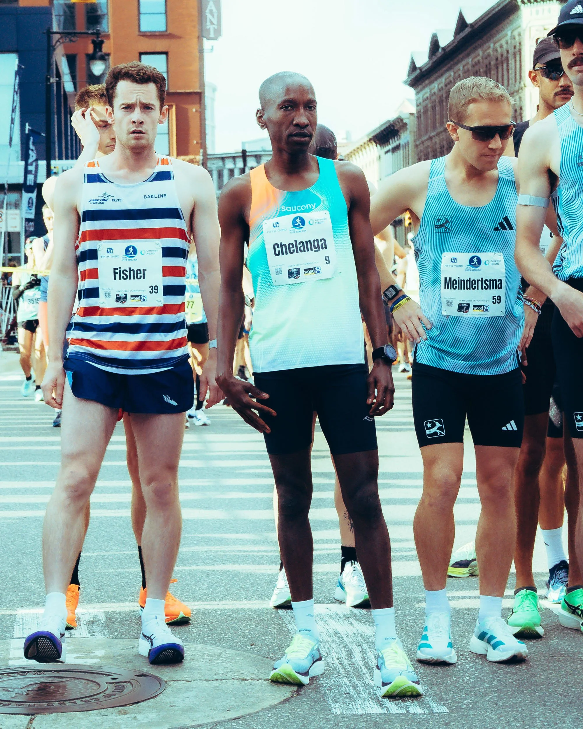A group of marathon runners standing at the start line in an urban street, dressed in athletic gear and sunglasses, with buildings in the background.