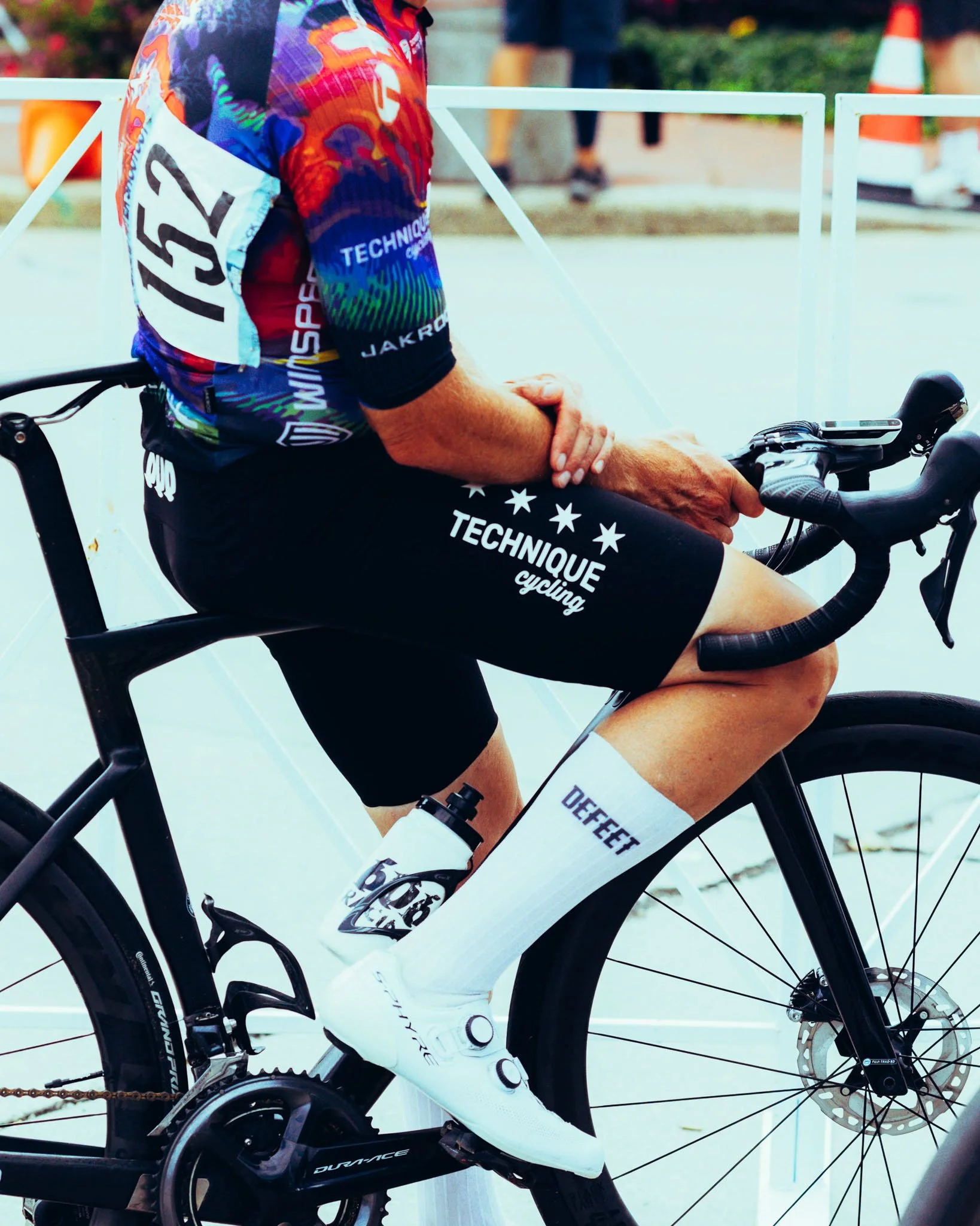 A person in cycling gear sitting on a white road bike, holding the handlebars, with a phone mounted on the handlebar. The cyclist is wearing a colorful jersey, black shorts with white text, and white socks with black logos. The background shows a blu