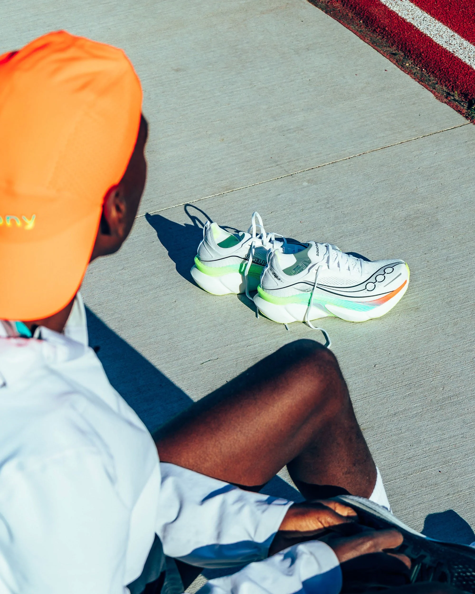 Person sitting on a sidewalk, wearing a white sports outfit with black and teal accents, a bright orange cap, and black shoes, with a pair of white and green running shoes placed on the ground nearby and a red textured curb in the background.