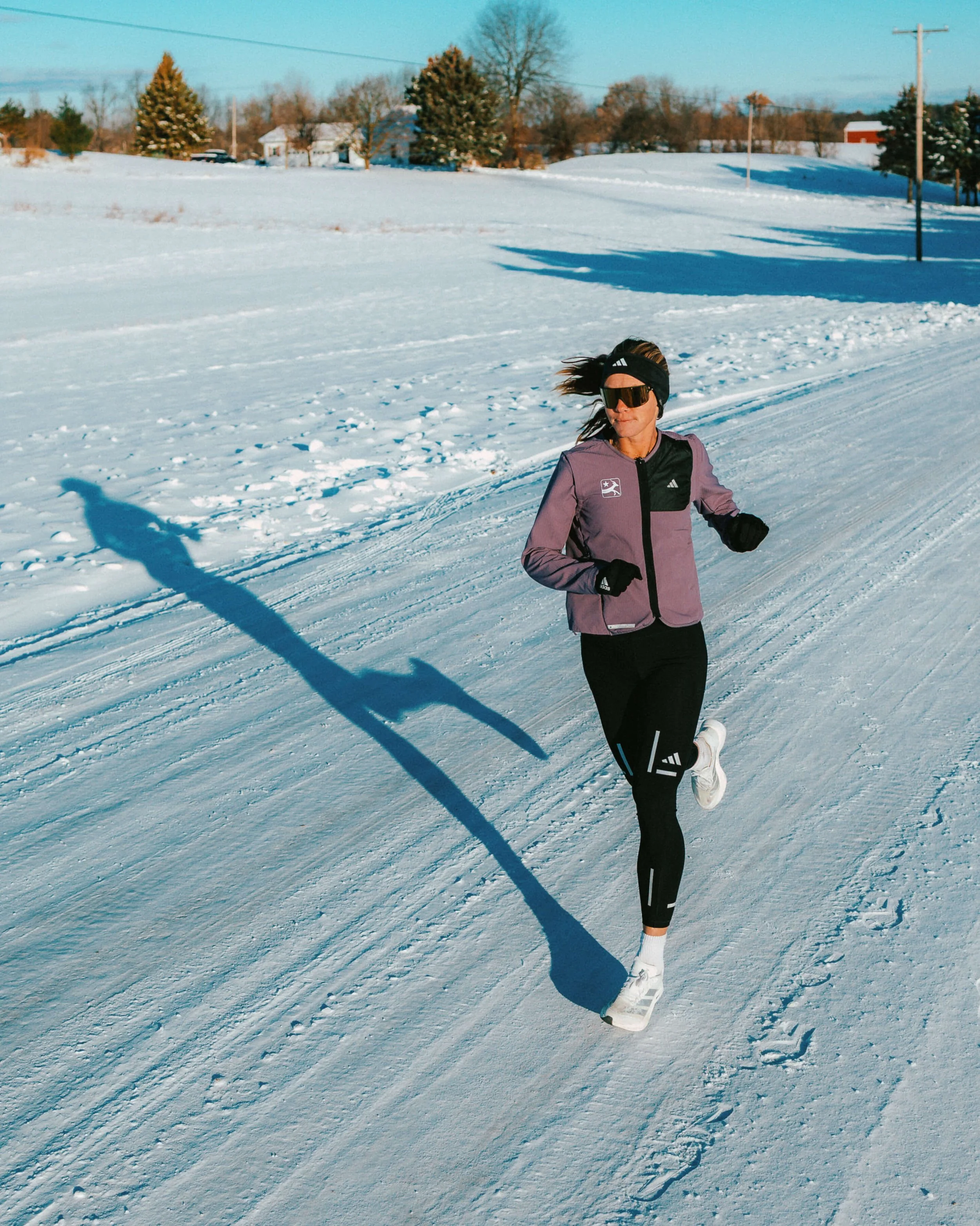 Woman running in snowy landscape wearing black running gear, sunglasses, and a headband, with houses and trees in the background.