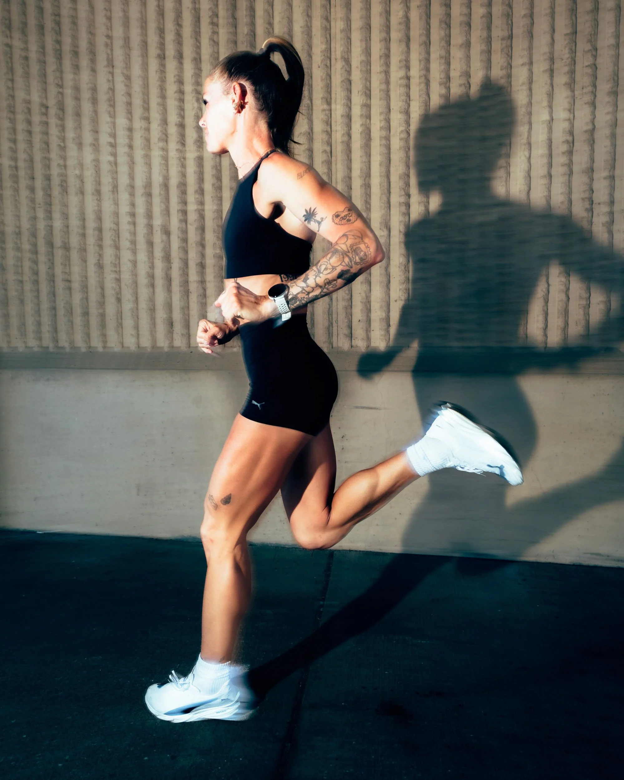 A woman with tattoos on her arm and side is running indoors against a textured wall. She is dressed in a black sports bra, black shorts, white socks, and sneakers, and is wearing a watch and earrings. Her hair is tied back in a ponytail, and her shad