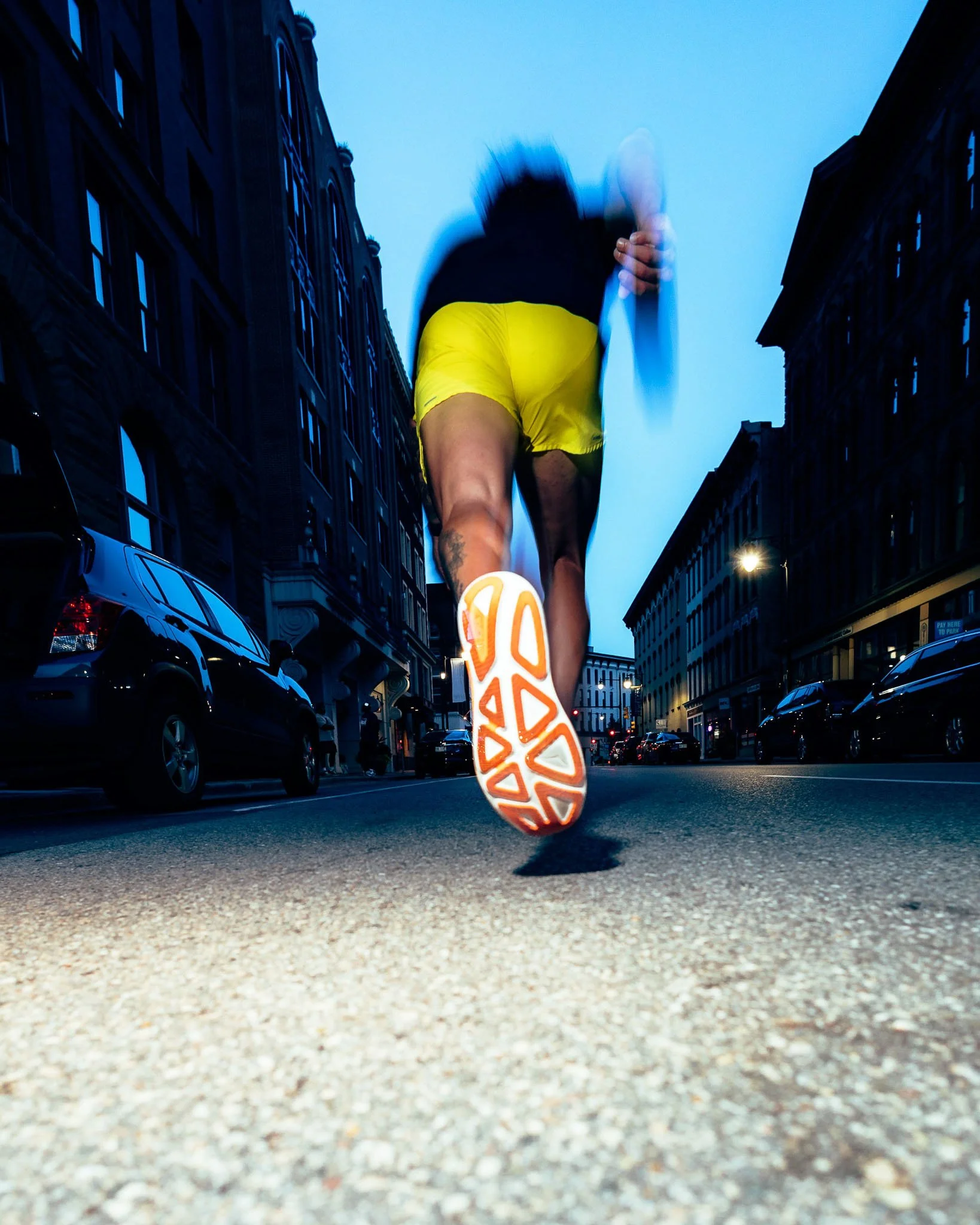 A person running on a city street at dusk, seen from a low angle, with the focus on their running shoes and legs. The person is wearing bright yellow shorts and a dark top, with their legs in mid-stride.