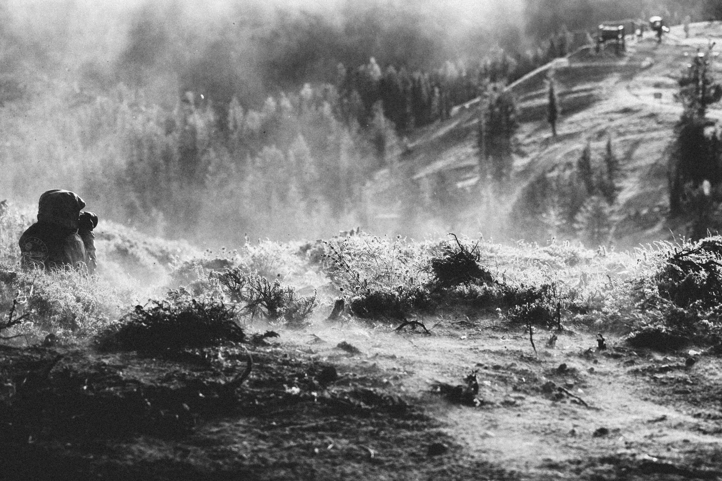 Black and white photo of soldiers walking through a forest with smoke and fire in the background.