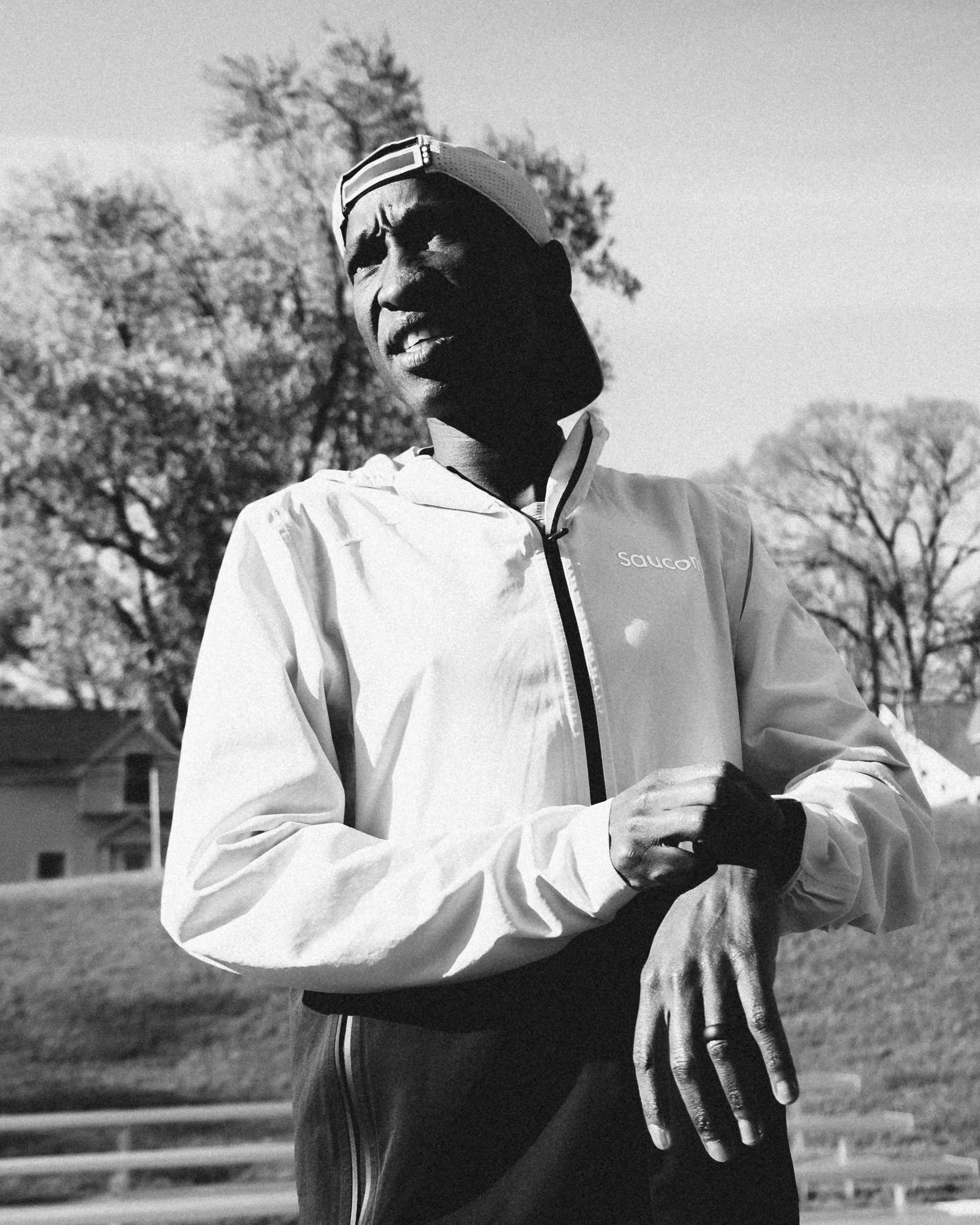 A person wearing a white jacket and a backwards cap, standing outdoors in a park with trees in the background, captured in black and white.