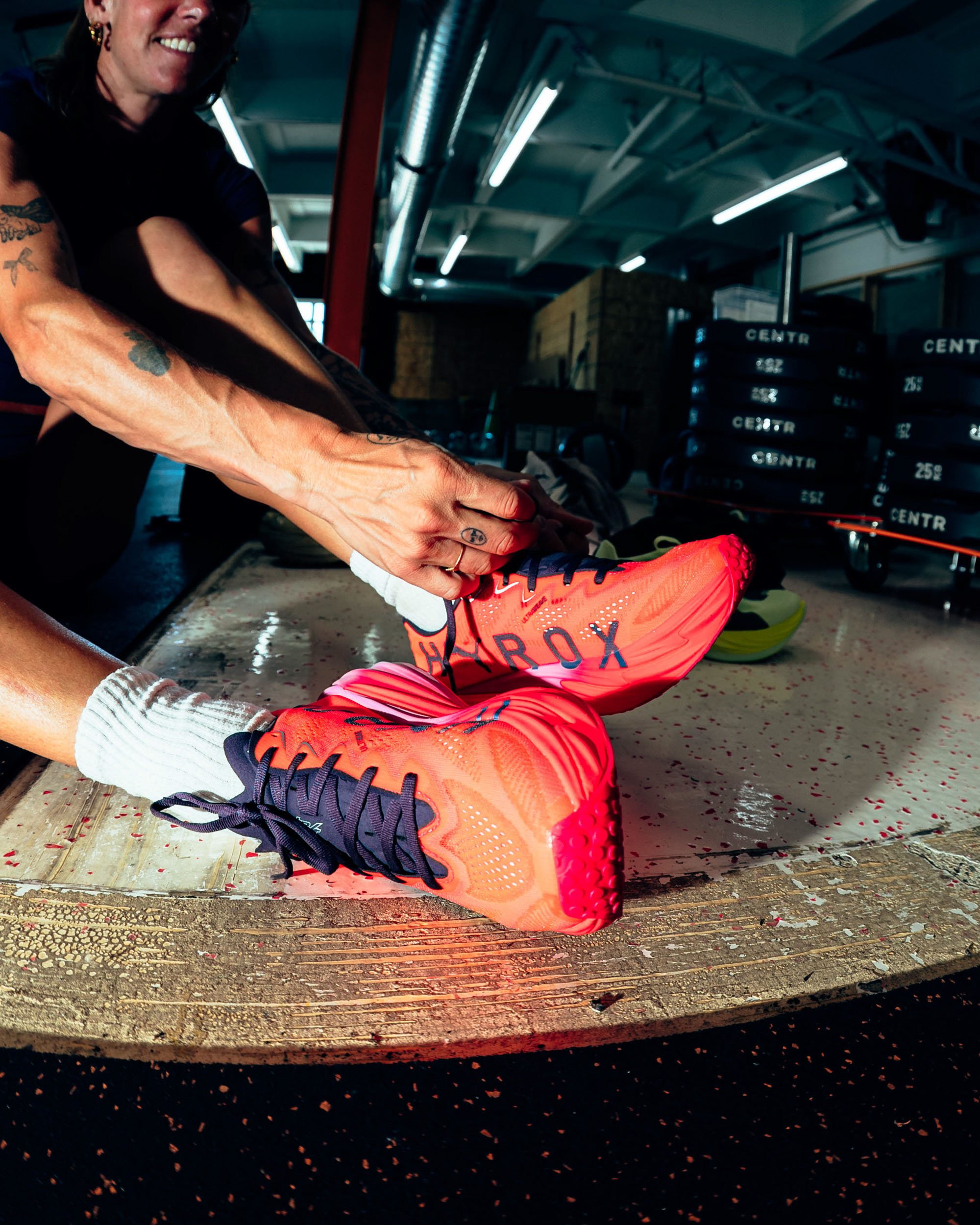 A person in athletic clothing and bright orange running shoes tying their shoelaces, seated on a wooden workout bench in a gym, with gym equipment and black rubber flooring visible in the background.