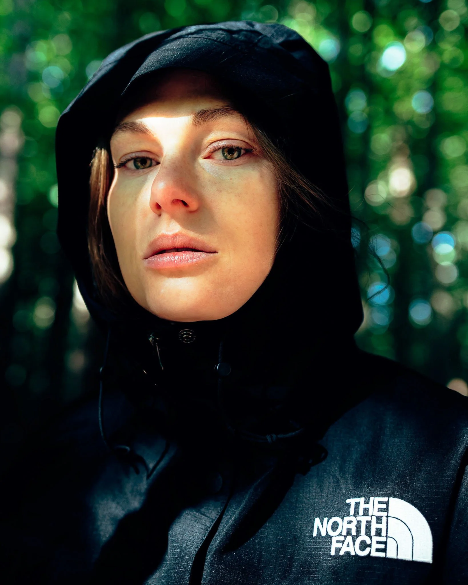 Close-up of a woman's face with green eyes, wearing a black hooded jacket with North Face logo, outdoors with blurred green foliage background.