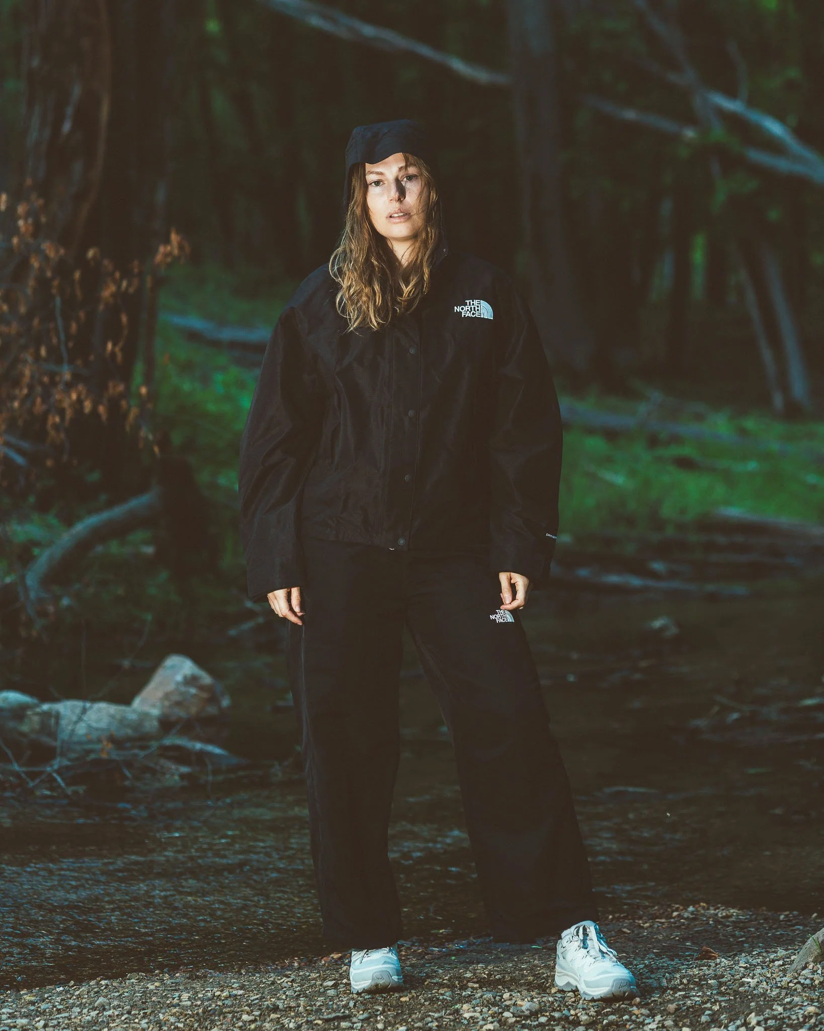 A woman standing on a rocky forest ground, wearing a black The North Face jacket and pants, white sneakers, and a black cap, in a dense wooded area.