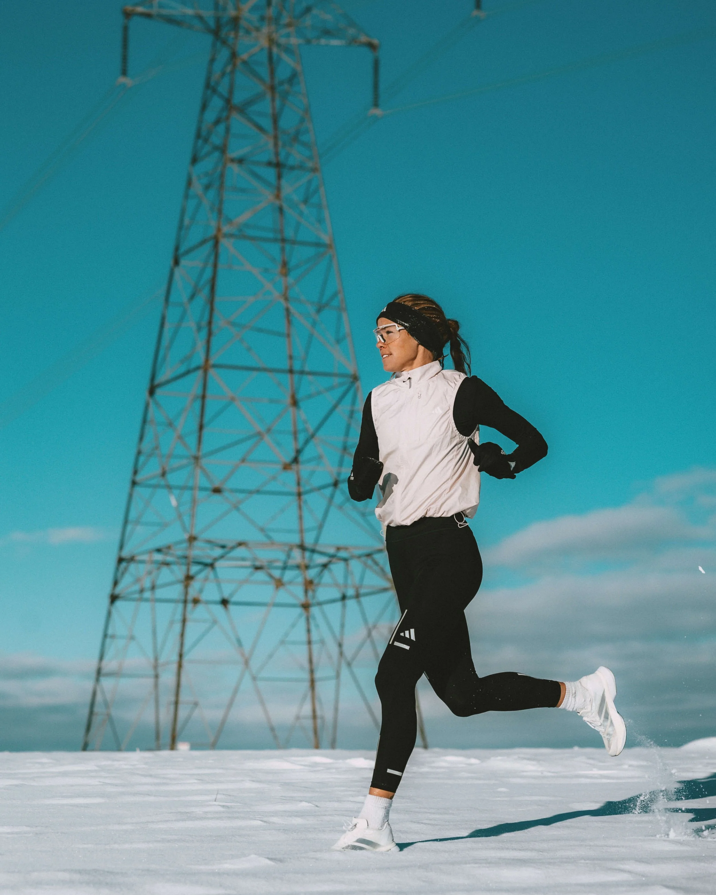 A woman running on snow near a large electricity pylon under a blue sky.