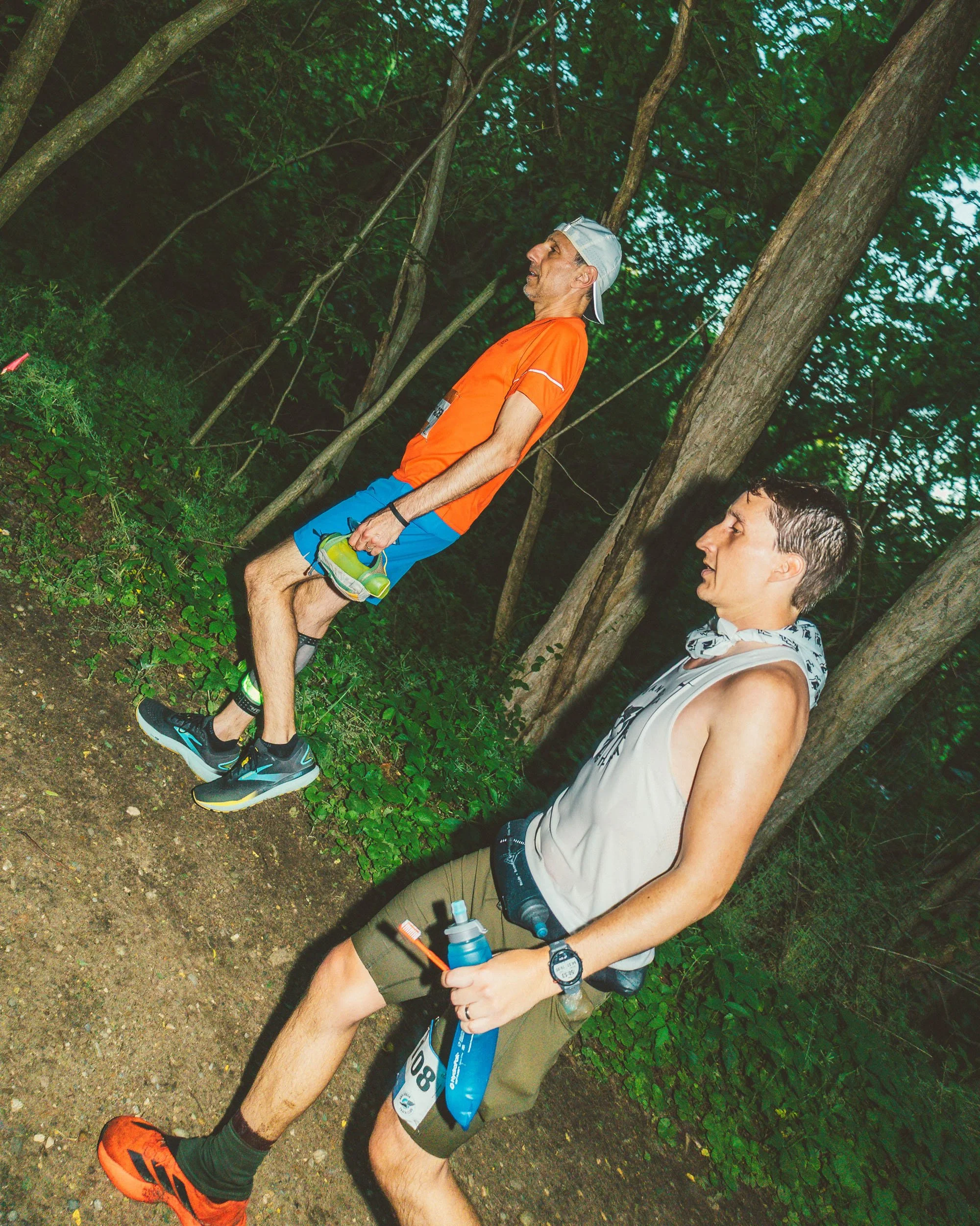Two male runners taking a break in a wooded area during a race, with one visibly holding water bottles and the other standing nearby, surrounded by trees and greenery.