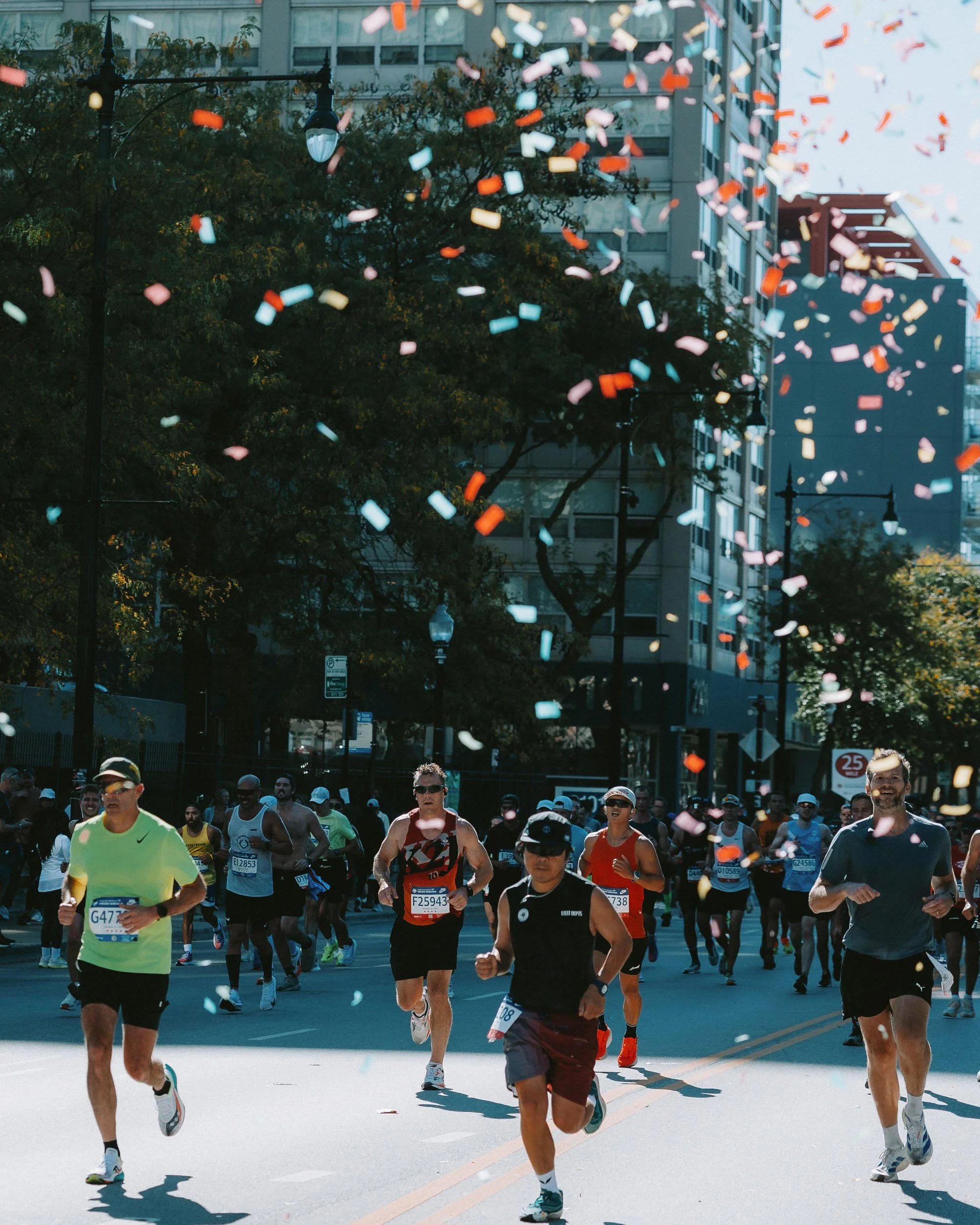 A large group of runners participate in a street race, with confetti falling from above and tall buildings and trees in the background.