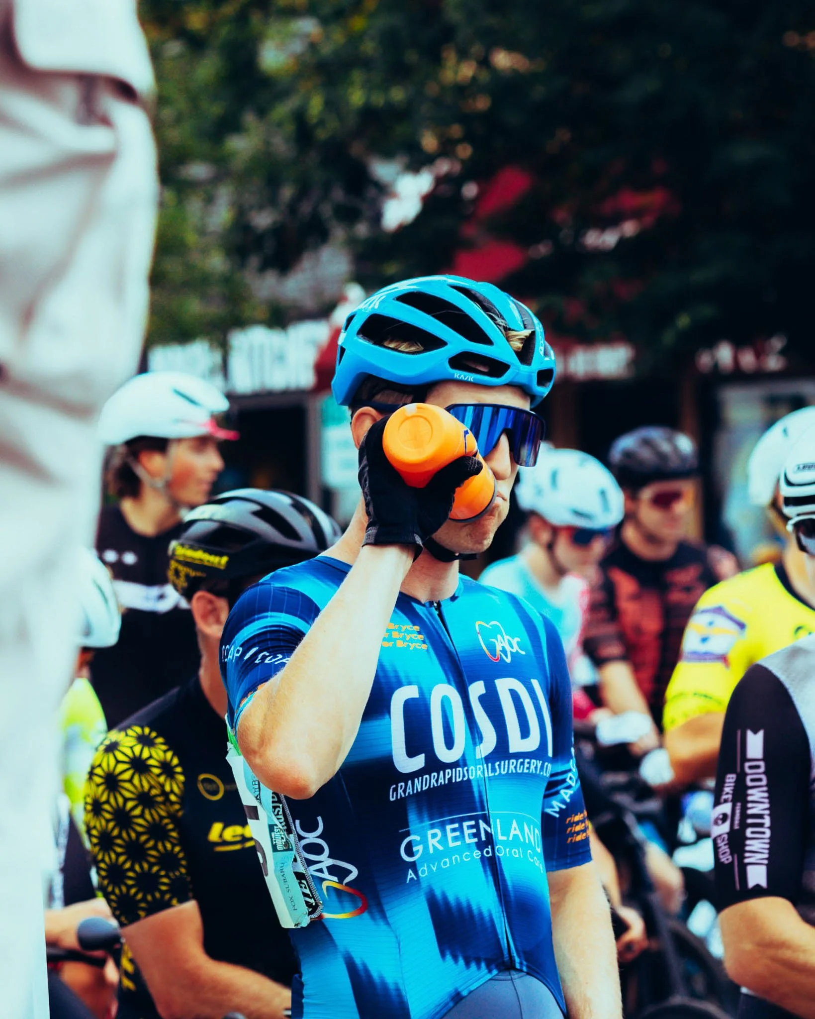 A cyclist wearing a blue helmet and sunglasses adjusting their helmet in a crowd of other cyclists, with trees and buildings in the background.