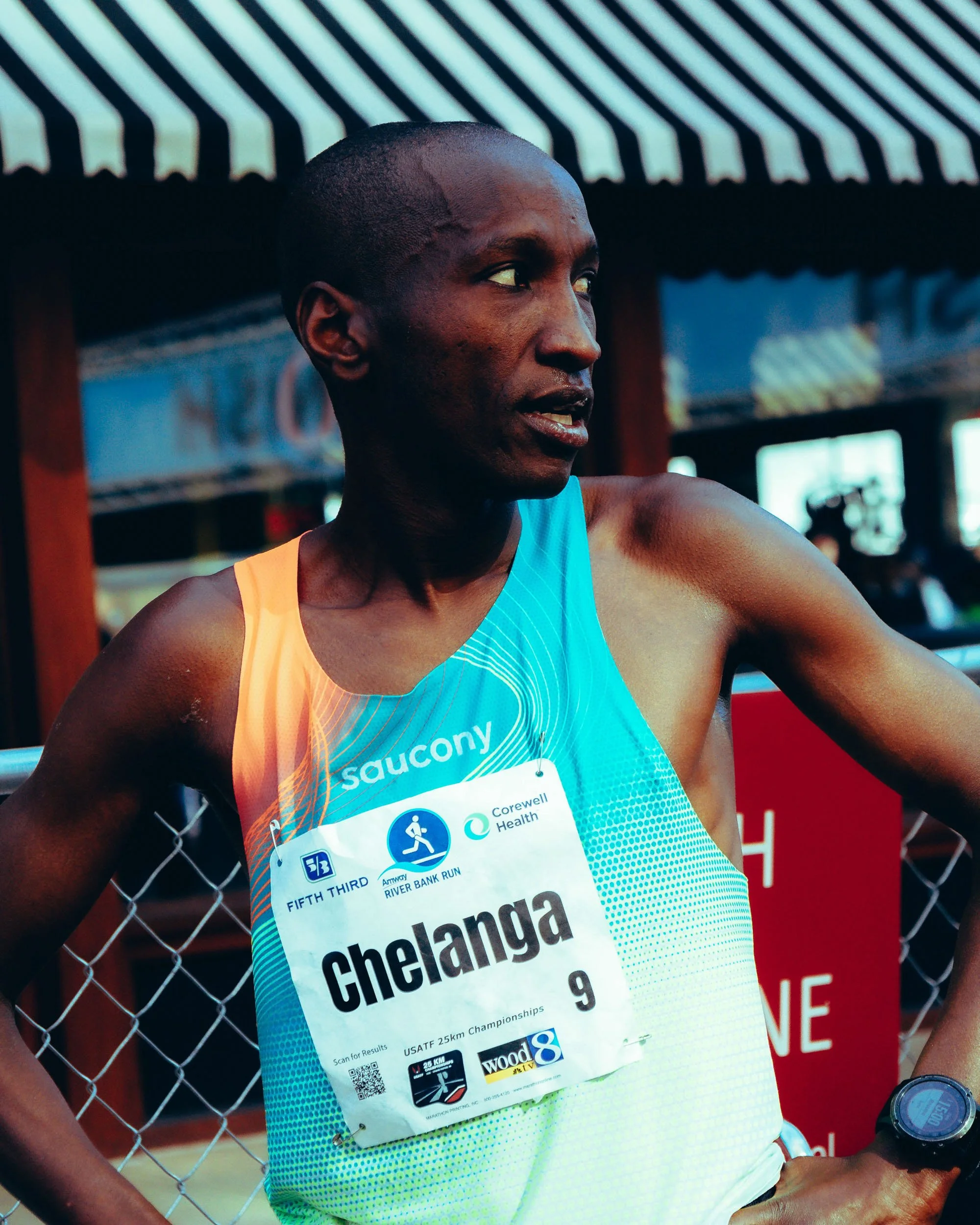 A male runner wearing a blue and orange athletic top, standing next to a chain link fence, with a race bib on his chest that reads 'chelanga' and indicates participation in a running event, with a race setup and booth in the background.