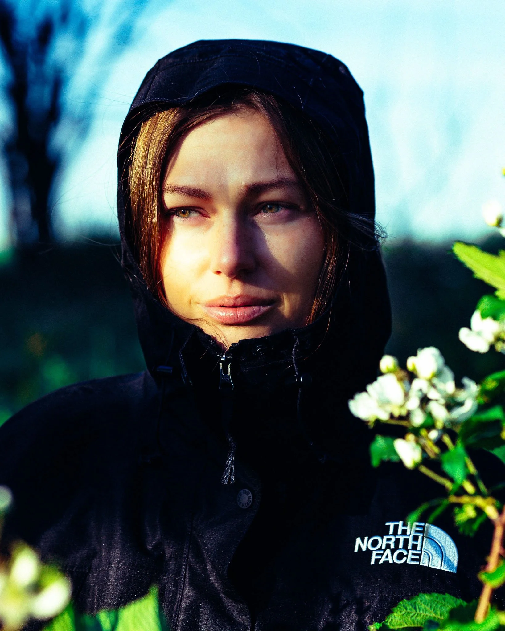 Close-up photo of a young woman with light brown hair, wearing a black hooded North Face jacket, standing outdoors amidst green foliage and white blooming flowers, with a blurred background.