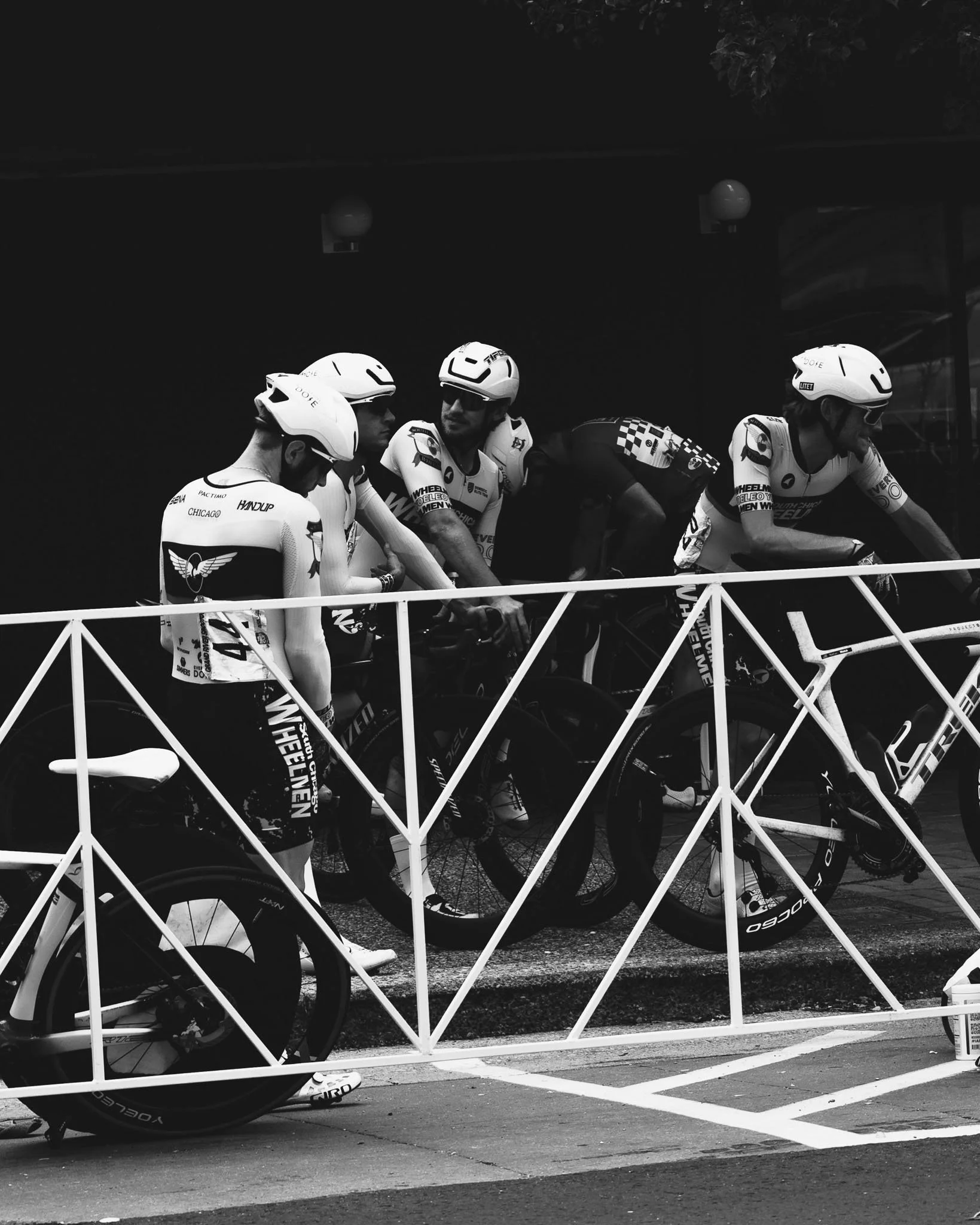 Black and white photo of a group of cyclist race riders wearing helmets and sports gear, gathering behind a white metal barricade on the street.